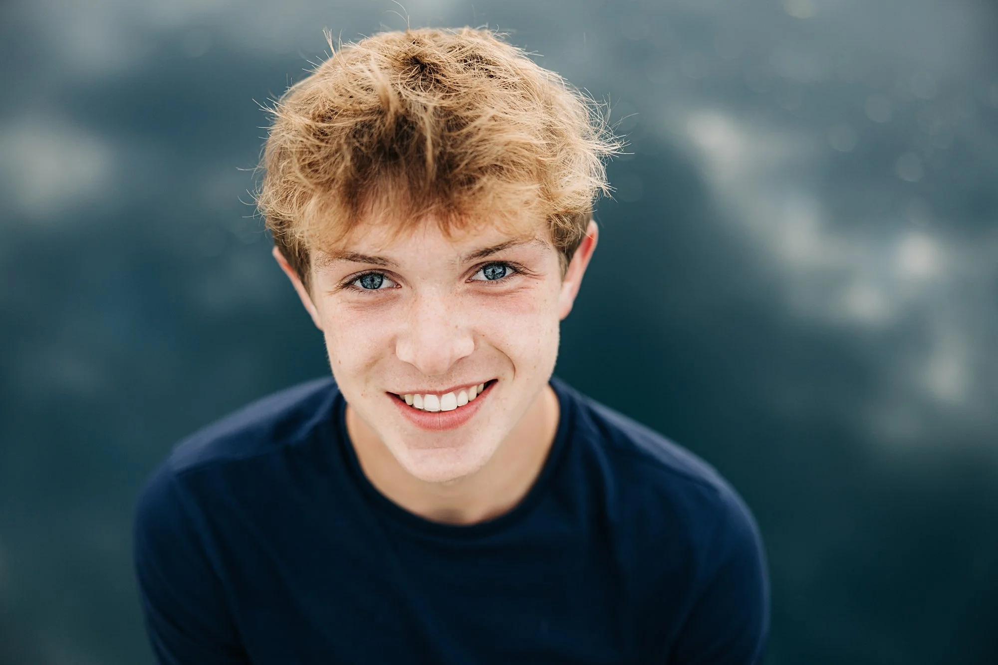 A young man with blond hair and blue eyes smiling at the camera, wearing a navy blue shirt, against a blurred dark background.