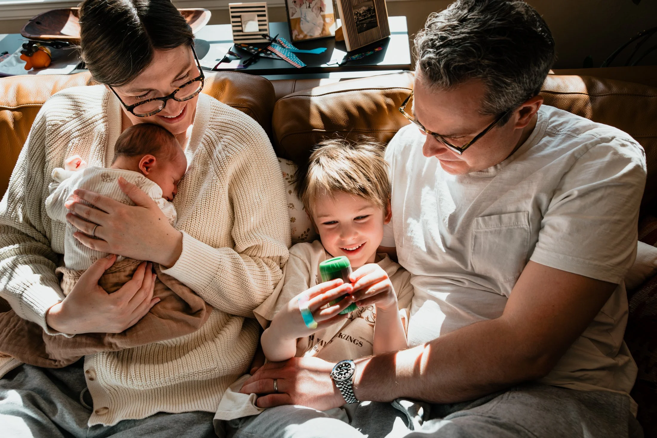 A happy family of four sitting on a leather couch, with a woman holding a newborn baby, and a boy holding a game controller, all smiling and enjoying their time together.