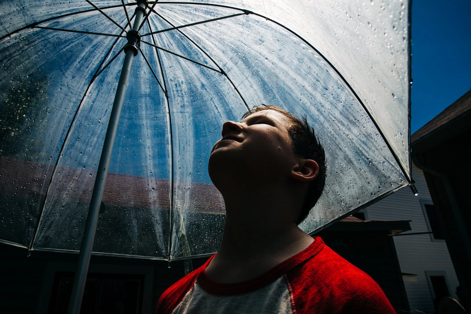 A boy holding a transparent umbrella with rain droplets, standing outdoors on a sunny day with a blue sky.