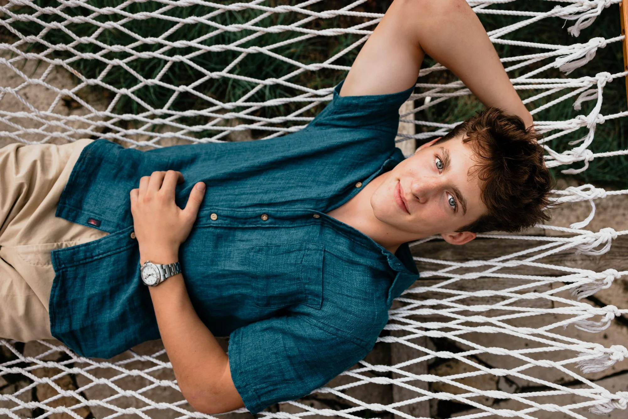 Young man lying in a hammock outdoors, wearing a blue shirt, khaki shorts, and a watch, resting with one arm behind his head and looking at the camera.