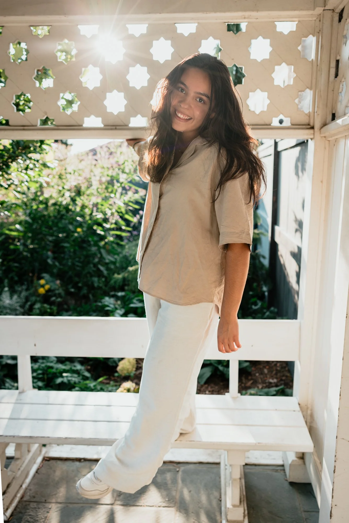 A woman smiling and looking at the camera, standing on a bench on a porch with sunlight shining through a decorative wooden backdrop, with greenery in the background.