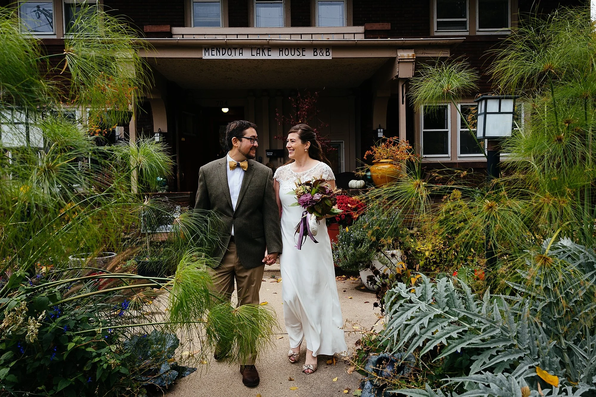 A couple in wedding attire holding hands and smiling at each other in front of a brown house with a sign that reads "Mendota Lake House B&B," surrounded by green plants and foliage.
