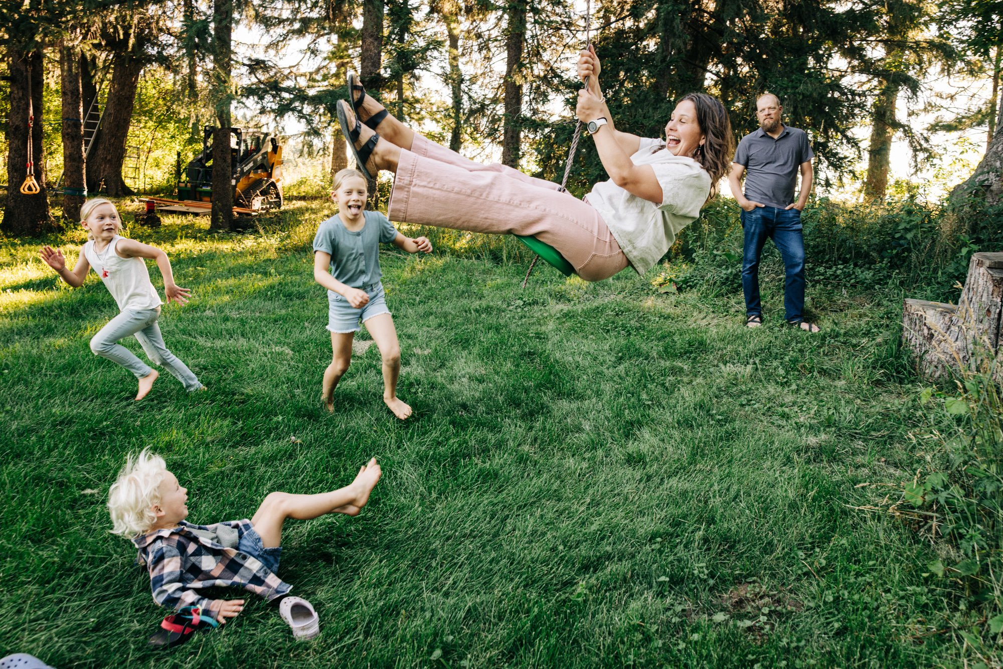 A woman on a swing laughs as she swings in the yard while four children play and run around on the grass, with a man standing in the background near trees.