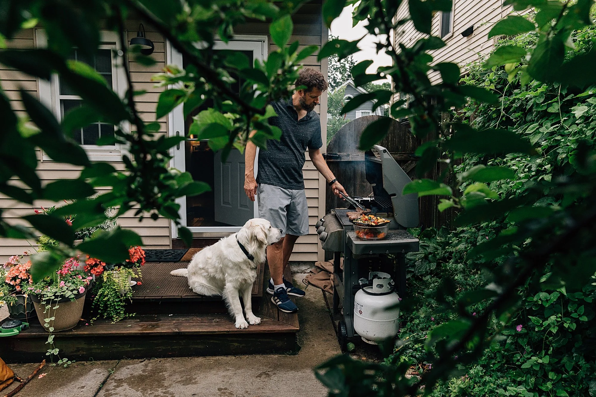 A man is cooking on a gas grill outside a house, with a golden retriever sitting nearby, surrounded by green foliage and potted plants.