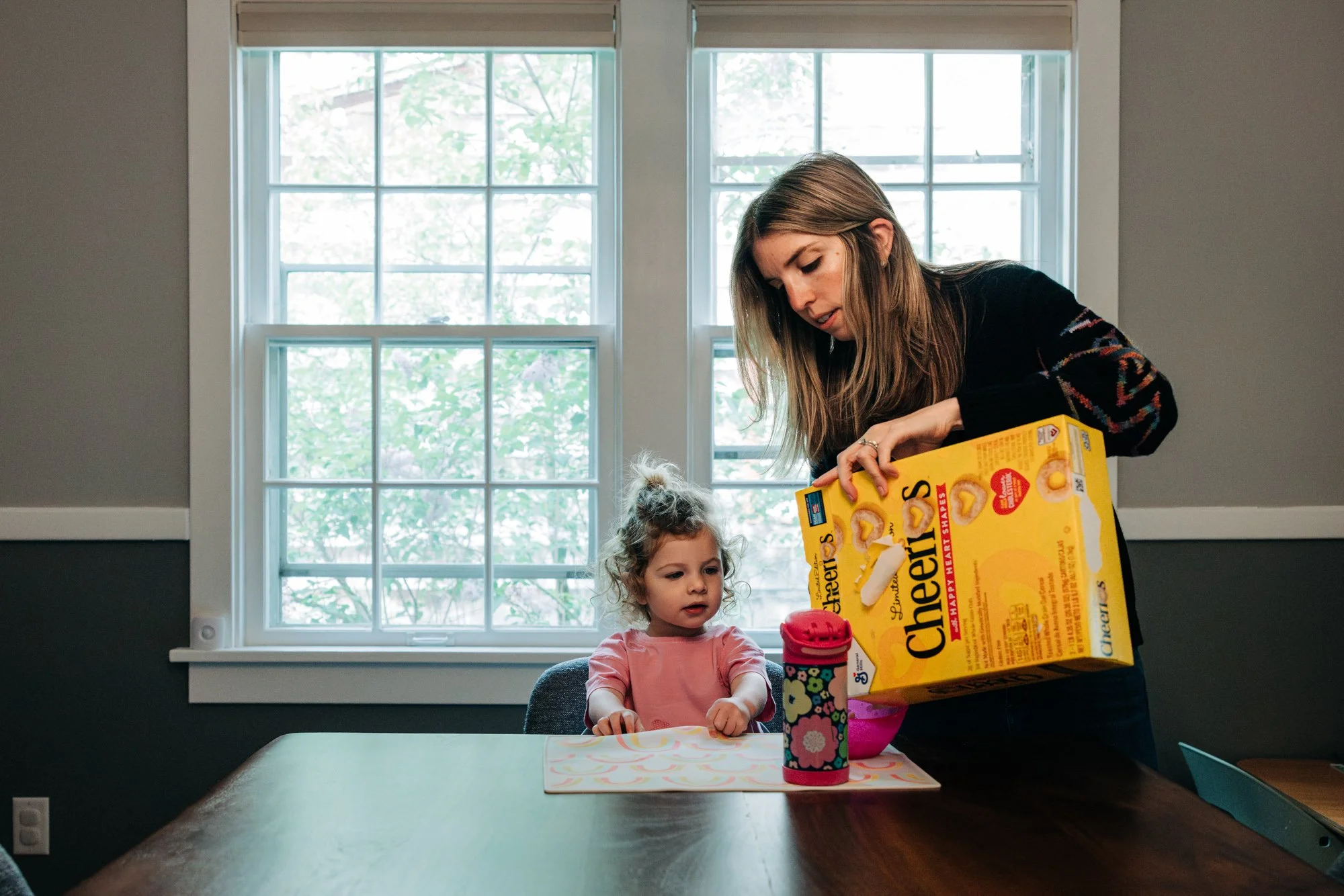 A woman pouring Cheerios cereal into a bowl for a young girl sitting at a dining table, with a window showing trees outside in the background.