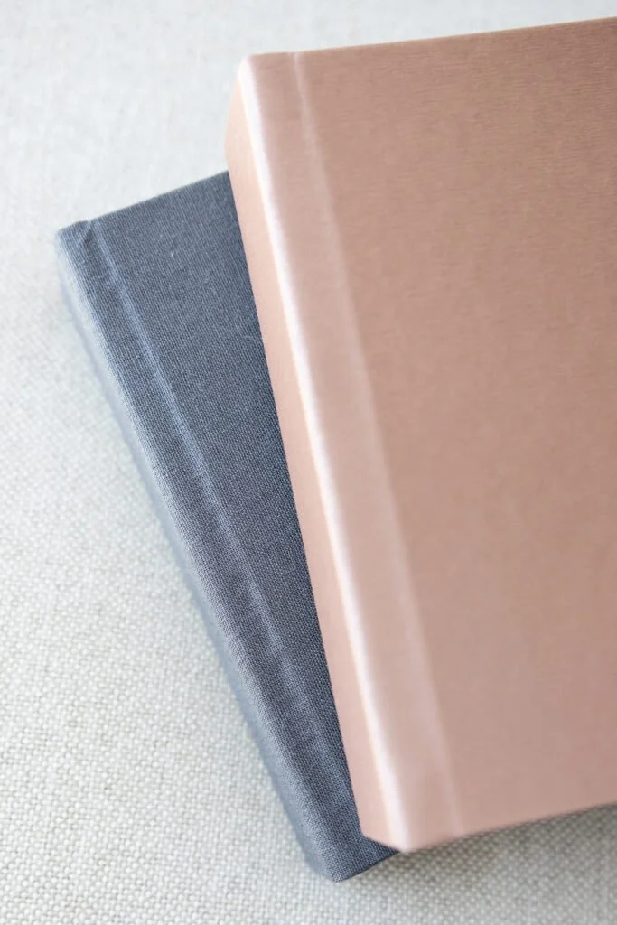 A close-up photograph of two closed notebooks stacked on top of each other on a light-colored textured surface. The top notebook has a pink cover and the bottom notebook has a dark gray cover.