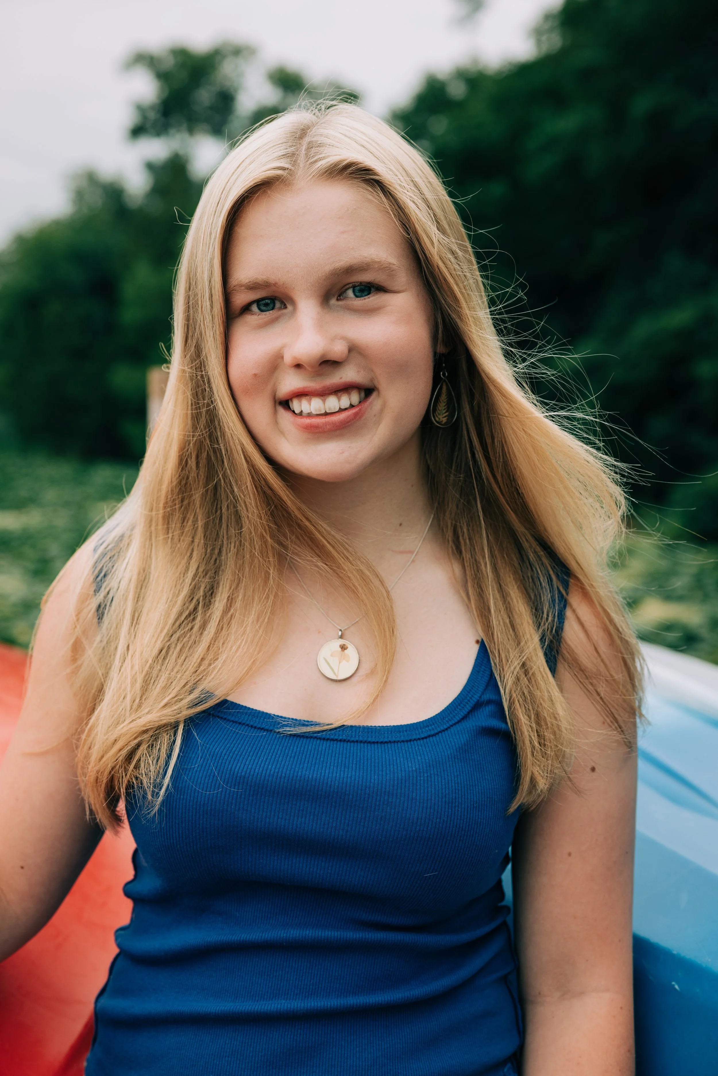 A young woman with blonde hair and blue eyes smiling outdoors, wearing a blue top, a necklace, and earrings, with a green, tree-lined background.