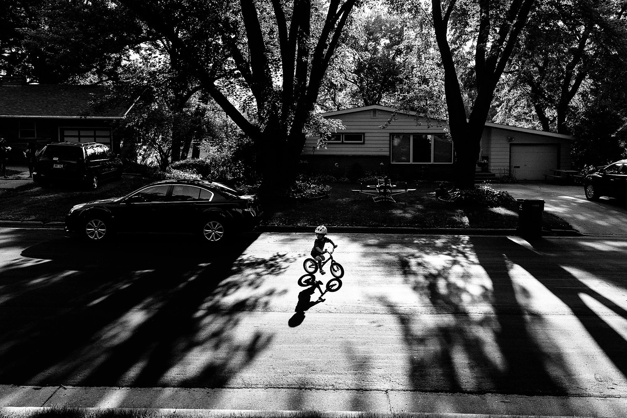 A young child riding a bicycle on a suburban street during daylight with shadows of trees cast on the pavement, black and white photograph.