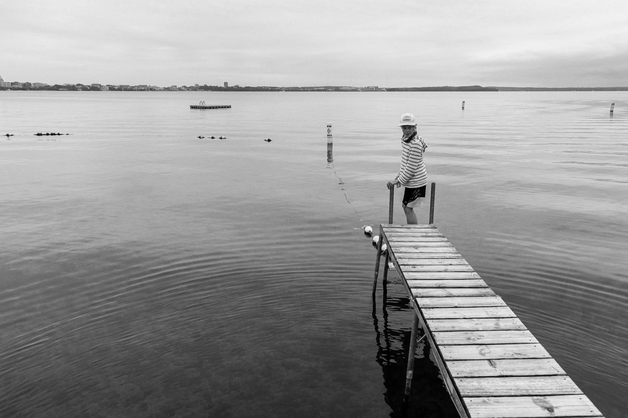 A boy in striped shirt and cap standing on a dock by the water, fishing with a line in a calm lake, with a city skyline in the distance.