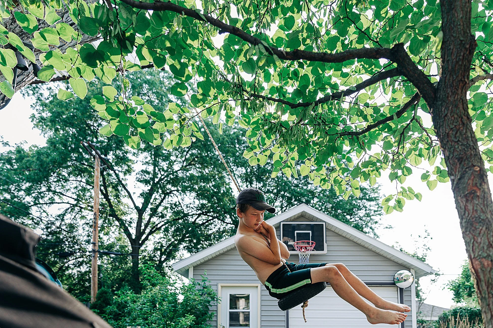 A shirtless boy in black shorts with green stripes, wearing a black cap, sitting on a tire swing hanging from a tree in a backyard, with trees, a house with gray siding, a basketball hoop, a satellite dish, and a chimney in the background.