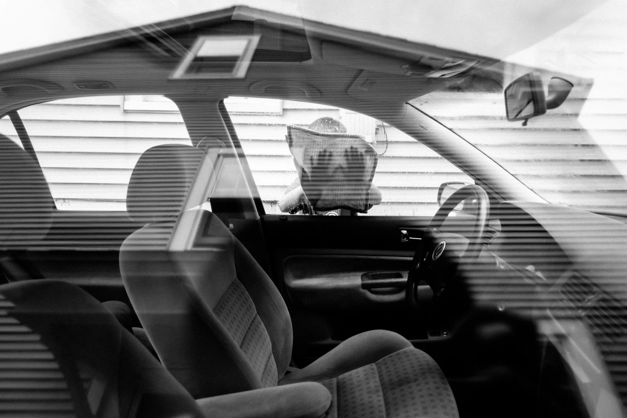 A black-and-white photo of the interior of a car, looking from outside through the windshield. The passenger seat is visible, along with a seatbelt. A person is seen outside the car, slightly blurred, taking the photo and wearing sunglasses.