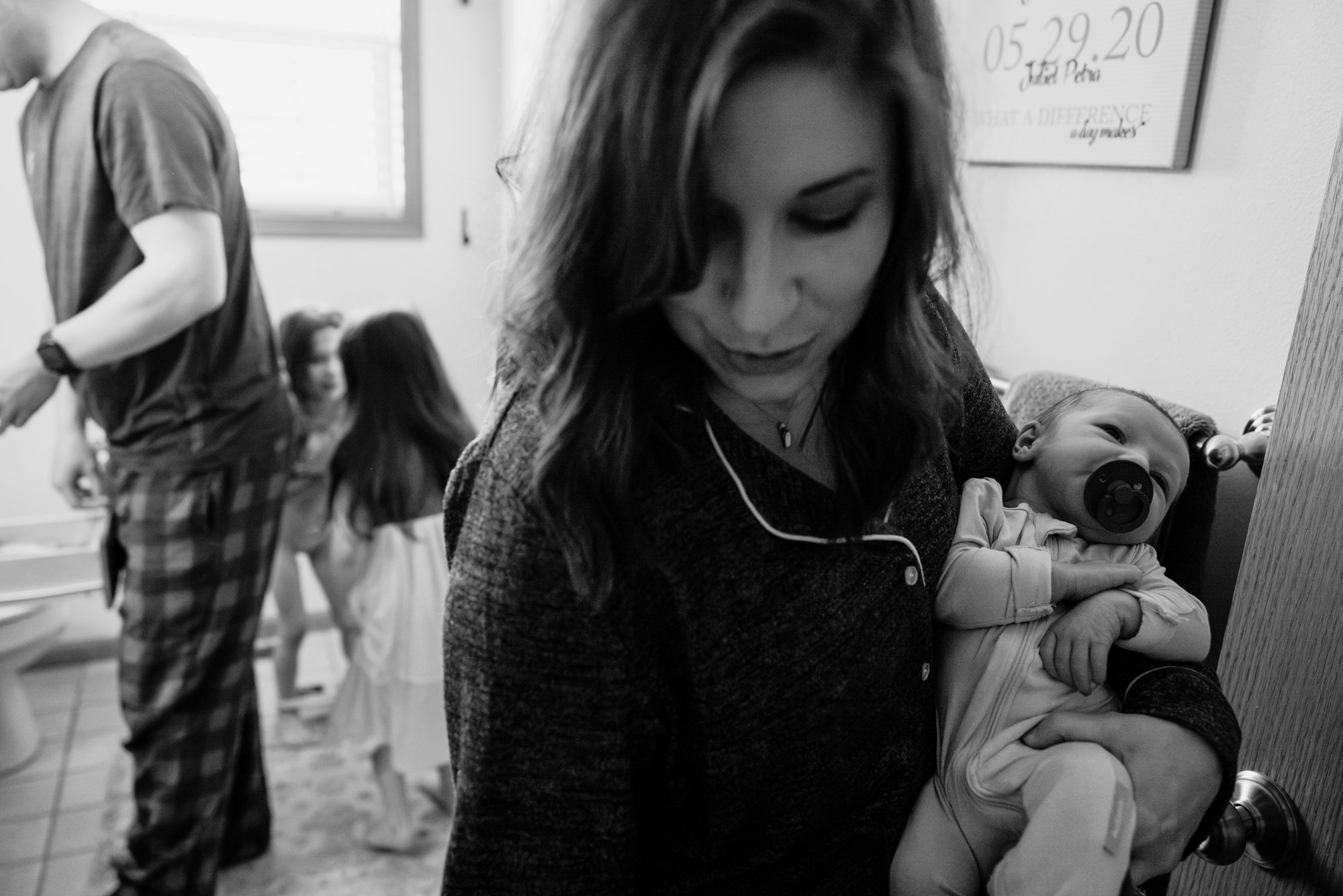 A woman holding a baby with a pacifier in their mouth, standing near a bathroom door. In the background, children are in the bathroom, with one sitting on the toilet and two standing nearby. The scene is indoors, in black and white.