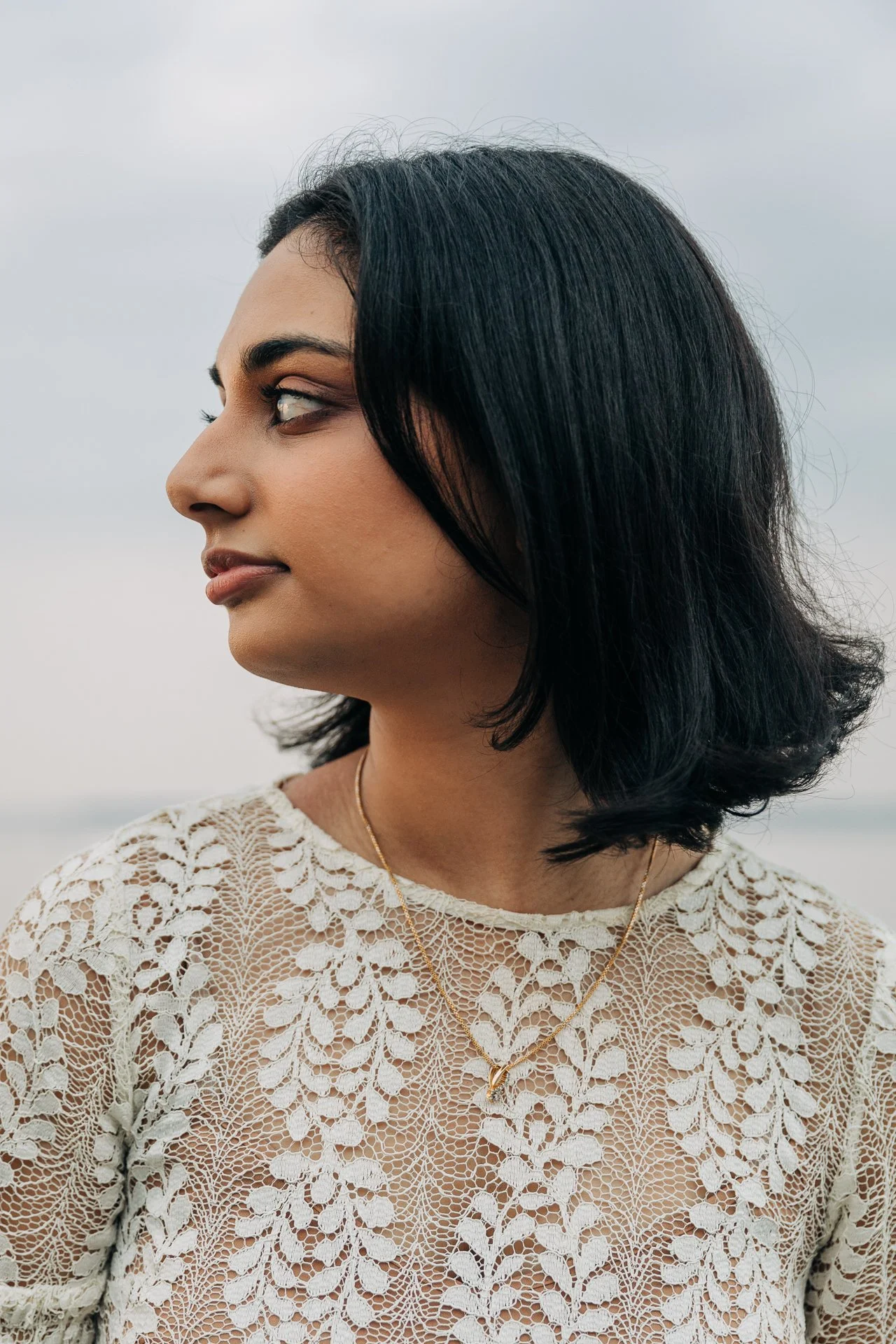 Close-up of a woman with black hair and tan skin, wearing a lace top and layered gold necklaces, looking to the side with a neutral expression, outdoors against a cloudy sky.
