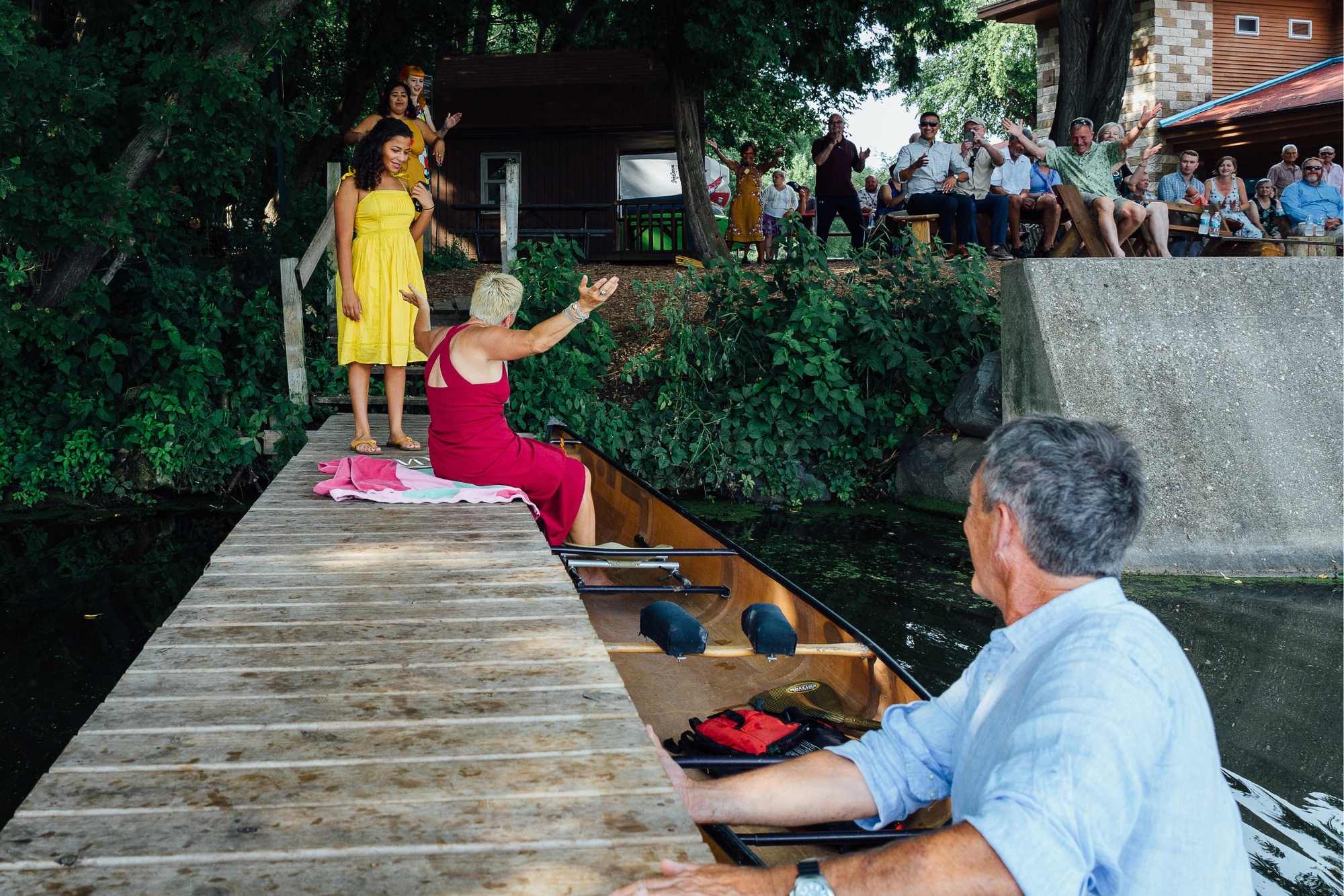 A group of people watching a woman in a red dress sit on a dock of a boat and raise her arms, while a young girl in a yellow dress stands nearby. The scene takes place outdoors near a lake with a wooded area and a deck with seated spectators in the b