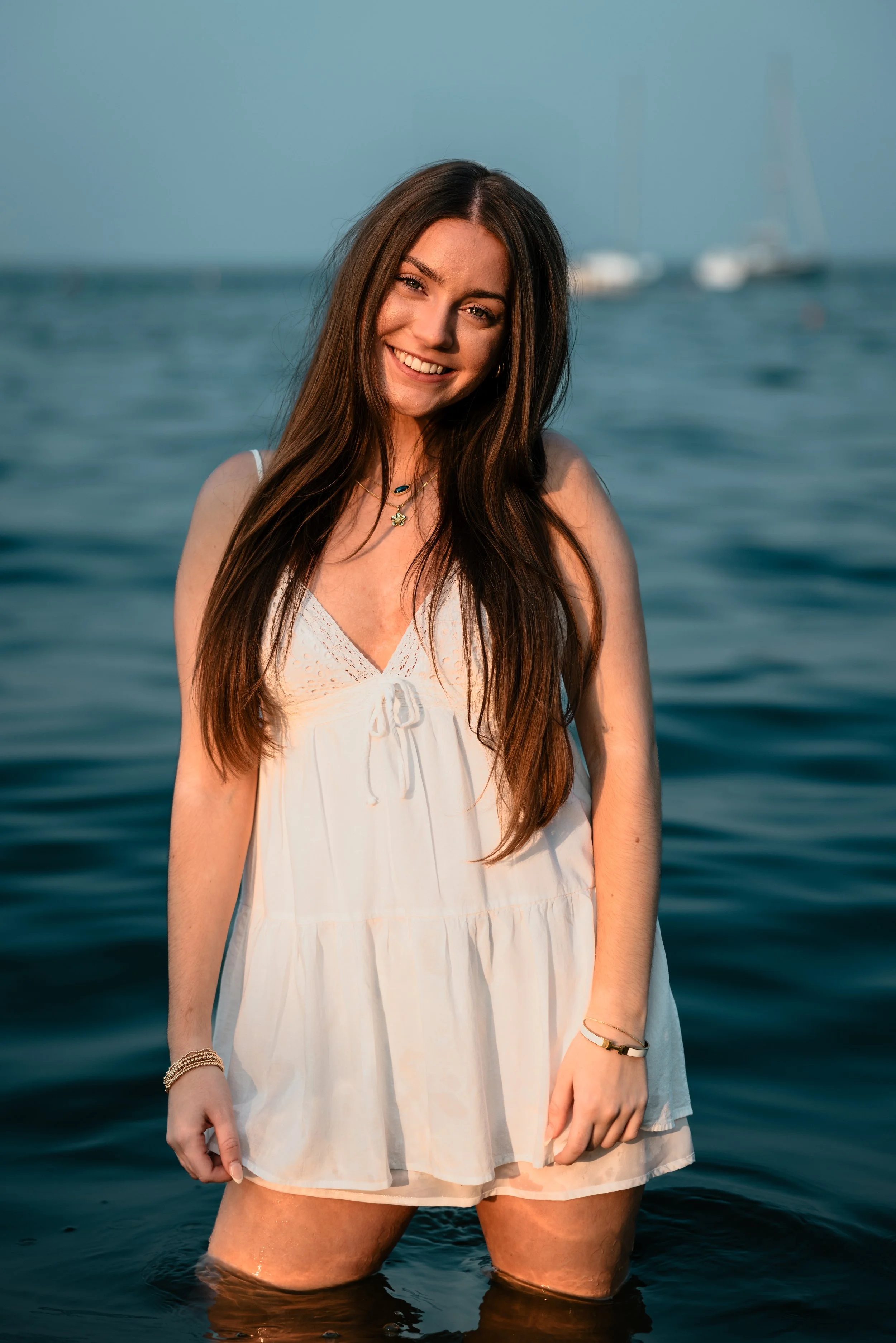 A young woman in a white dress standing in the water at the beach with sailboats in the background, smiling.