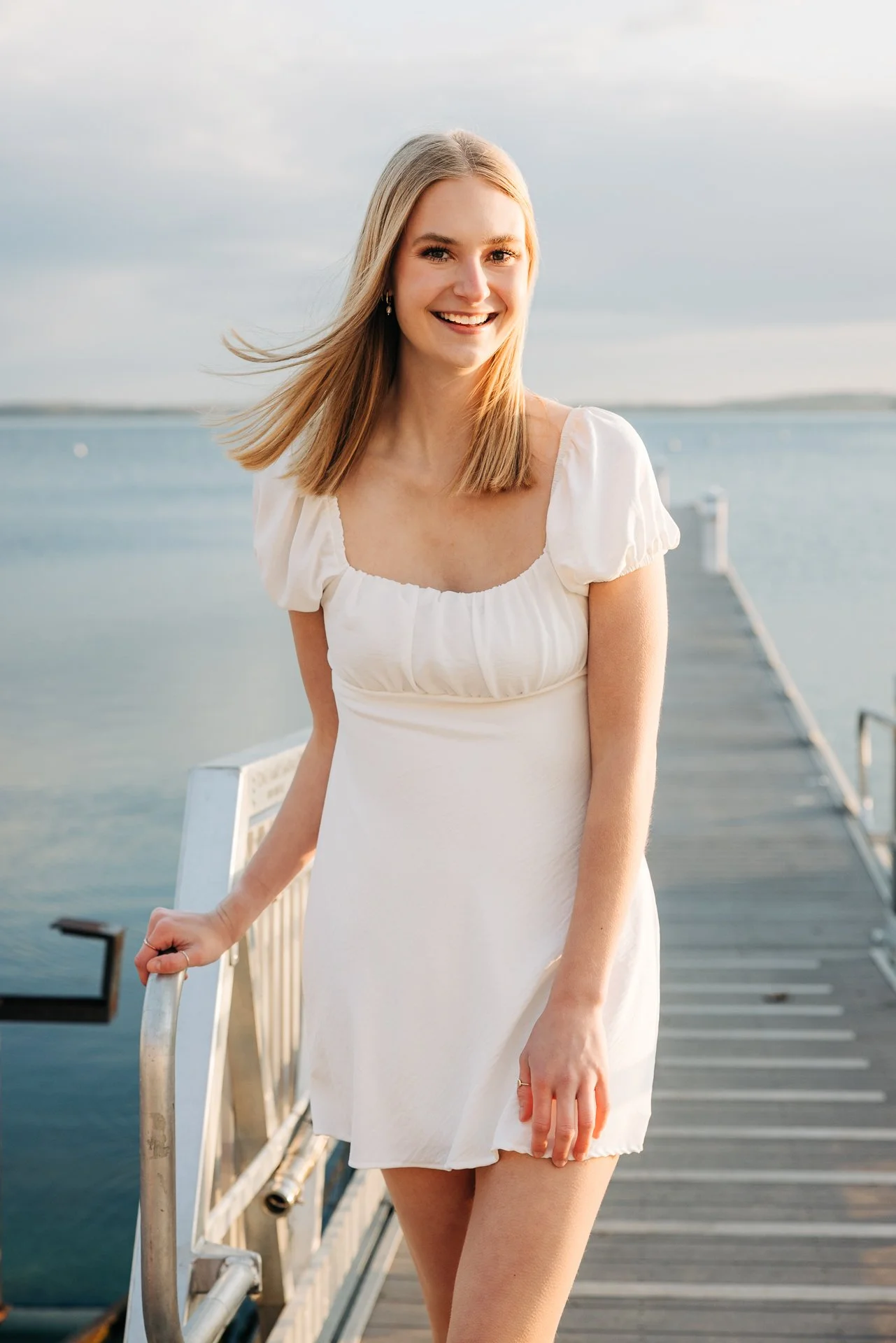 A young woman with blonde hair, wearing a white dress, standing on a pier by the water, smiling at the camera.