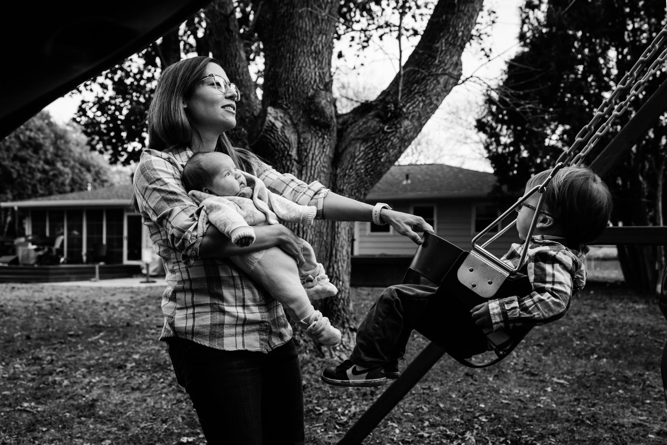 A woman with glasses holding a baby in her arms, standing next to a child in a swing, outdoors in a backyard with trees and houses in the background.