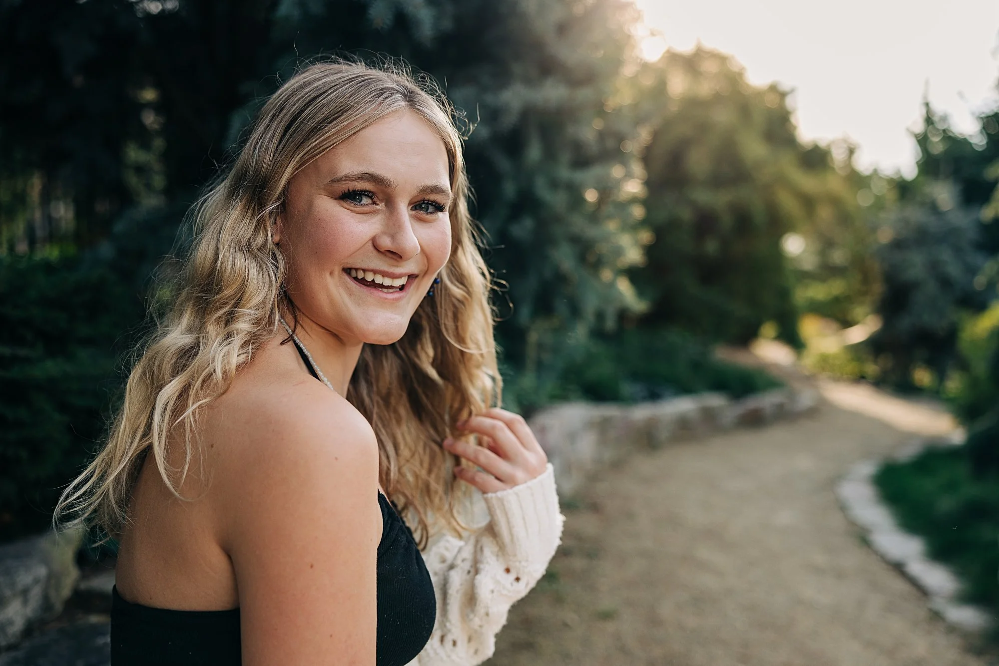 A young woman with long blonde hair, smiling, in an outdoor park setting during sunset.
