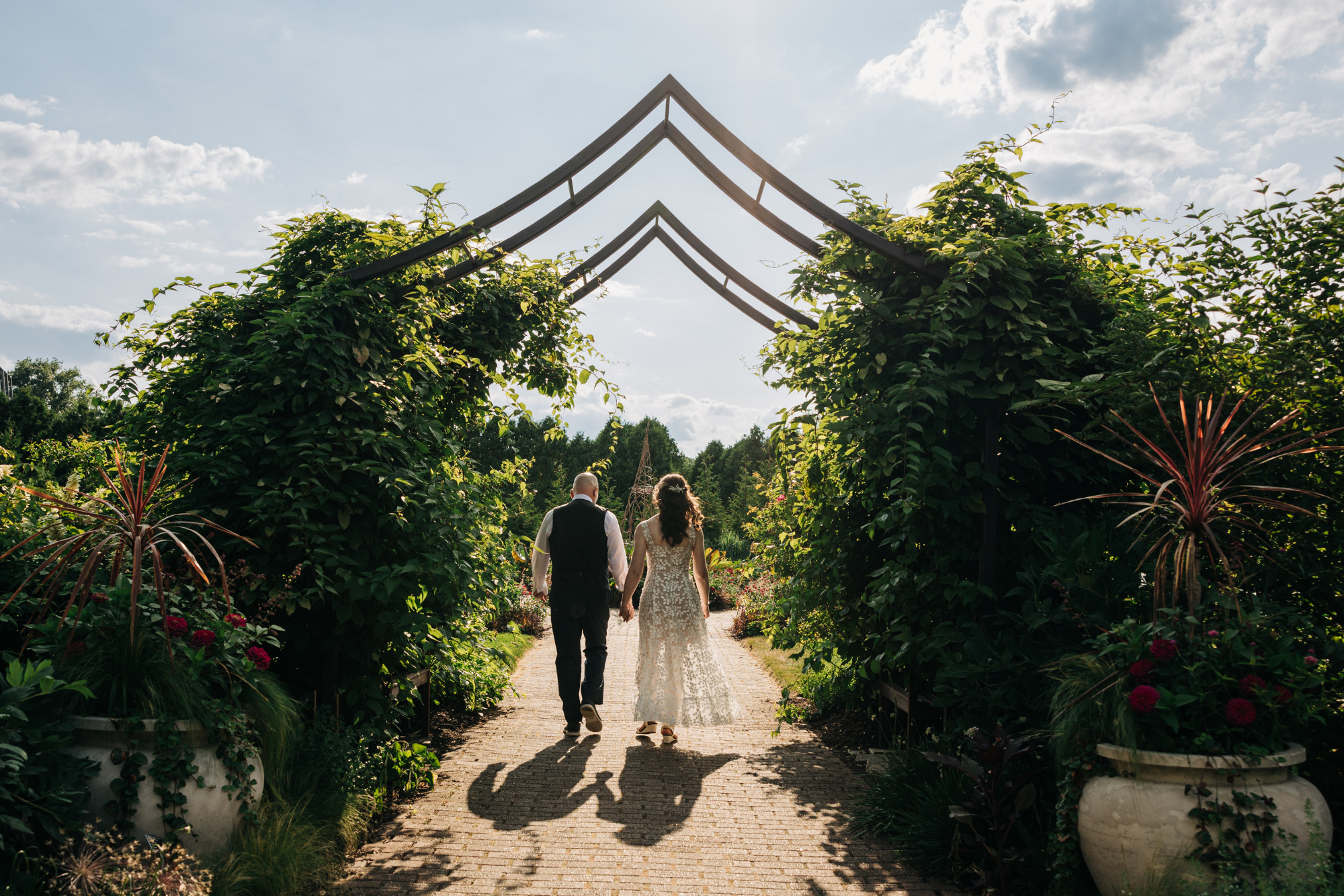 A couple holding hands walking through a garden archway on a sunny day.