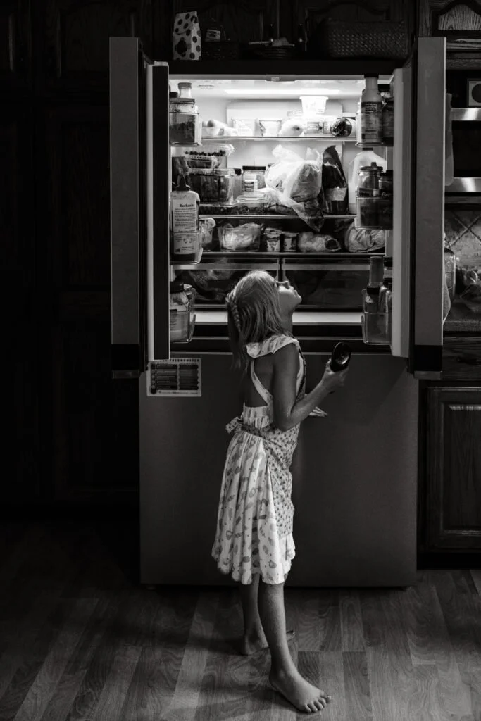 Young girl with shoulder-length hair, wearing a dress with a patterned half apron, standing barefoot in front of an open refrigerator, looking inside while holding a small container.
