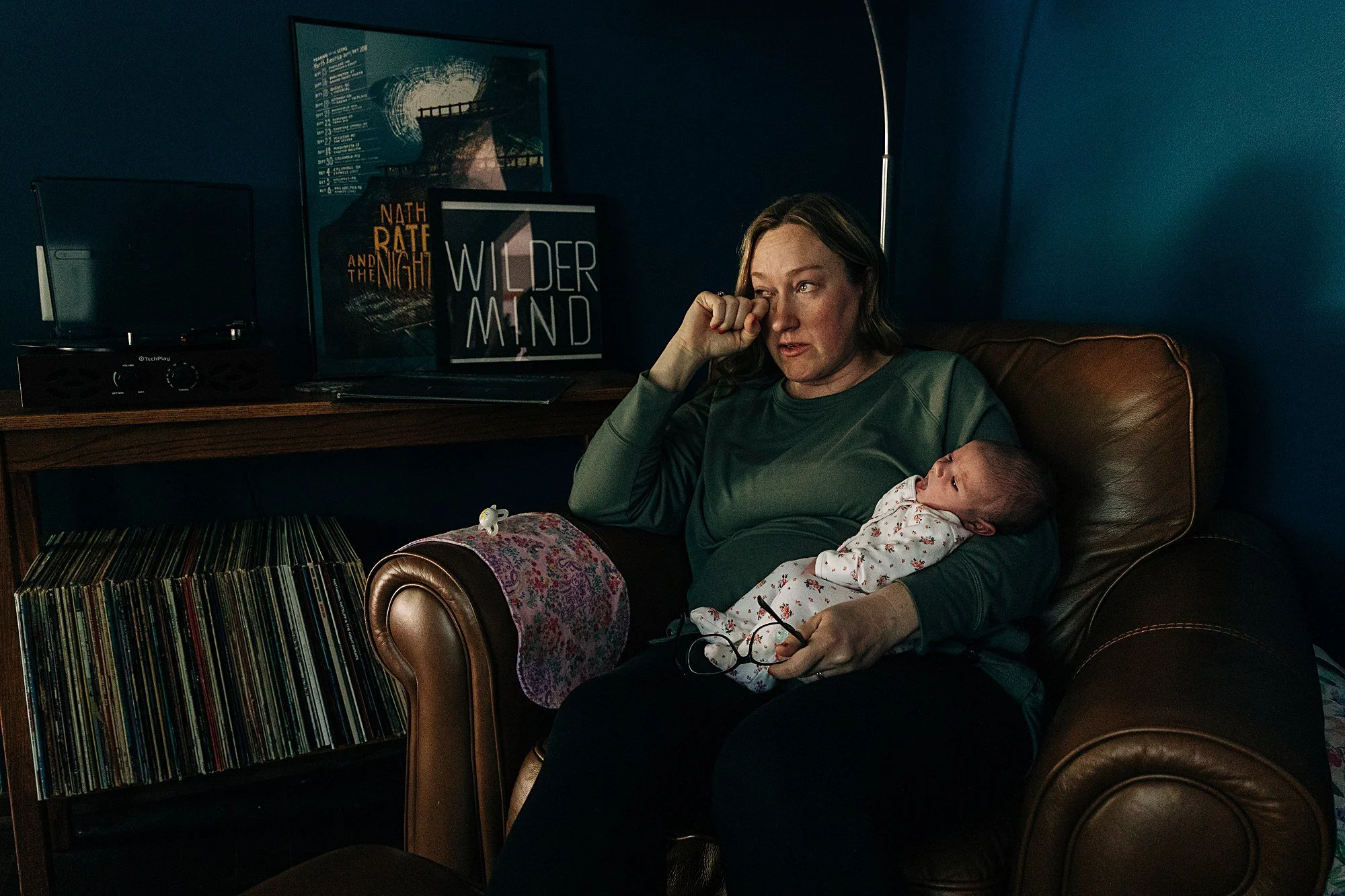 A woman sitting on a brown leather couch in a dark room, holding a baby in her lap. The woman is rubbing her eye with her hand and has glasses resting on her lap. The baby is dressed in a white outfit with red and black patterns. There is a record co