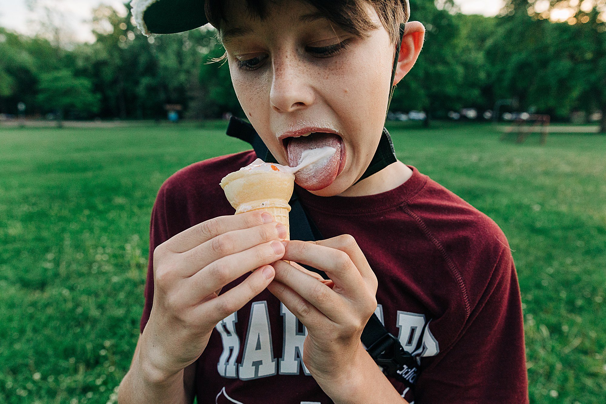 A young boy wearing a maroon shirt and a helmet, licking ice cream outdoors in a grassy park.