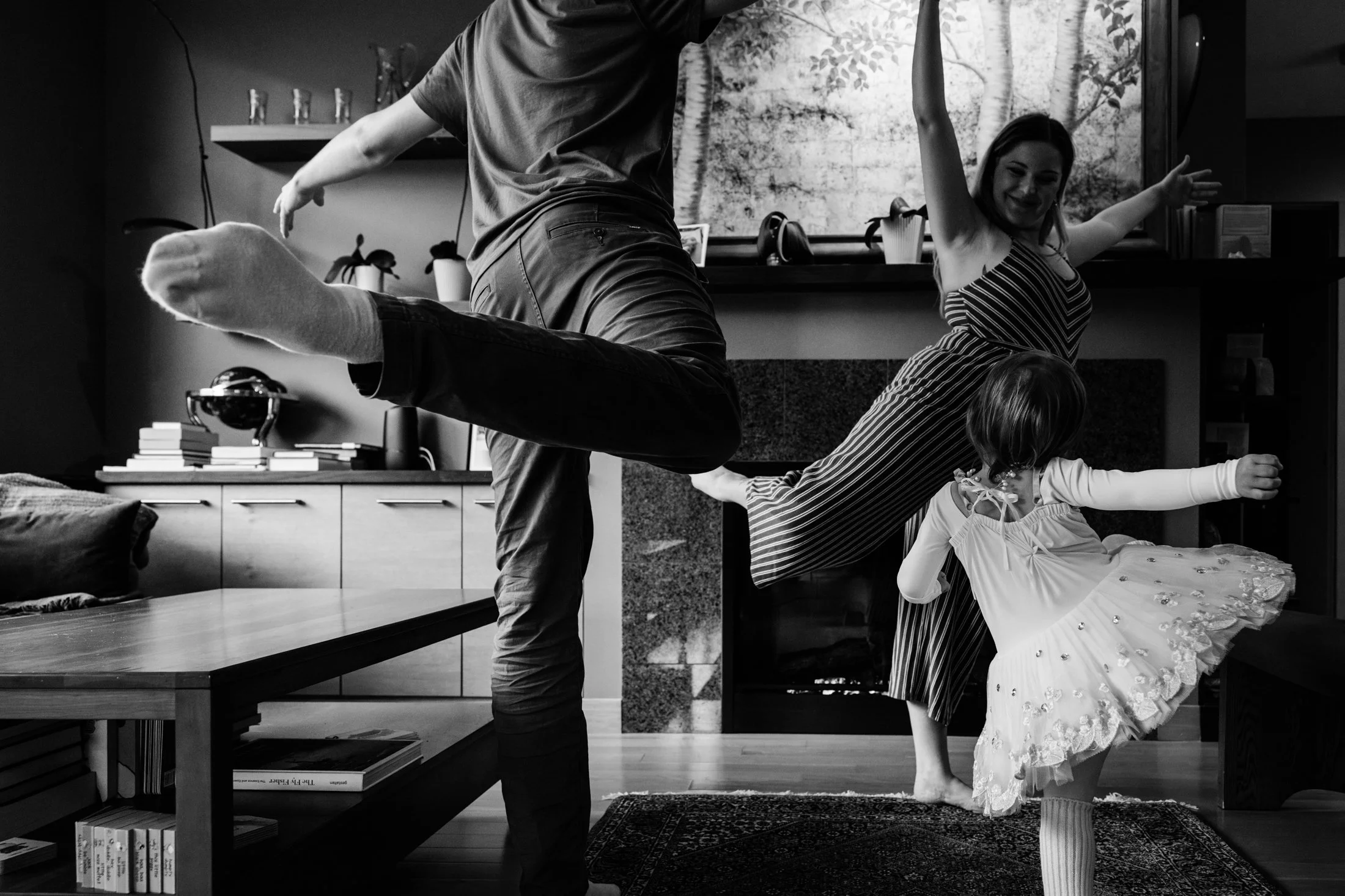 A family of four having fun indoors, with the father balancing on one leg, the mother smiling in a striped dress, and two young children, one in a dress and the other in pajamas, playing enthusiastically around them.