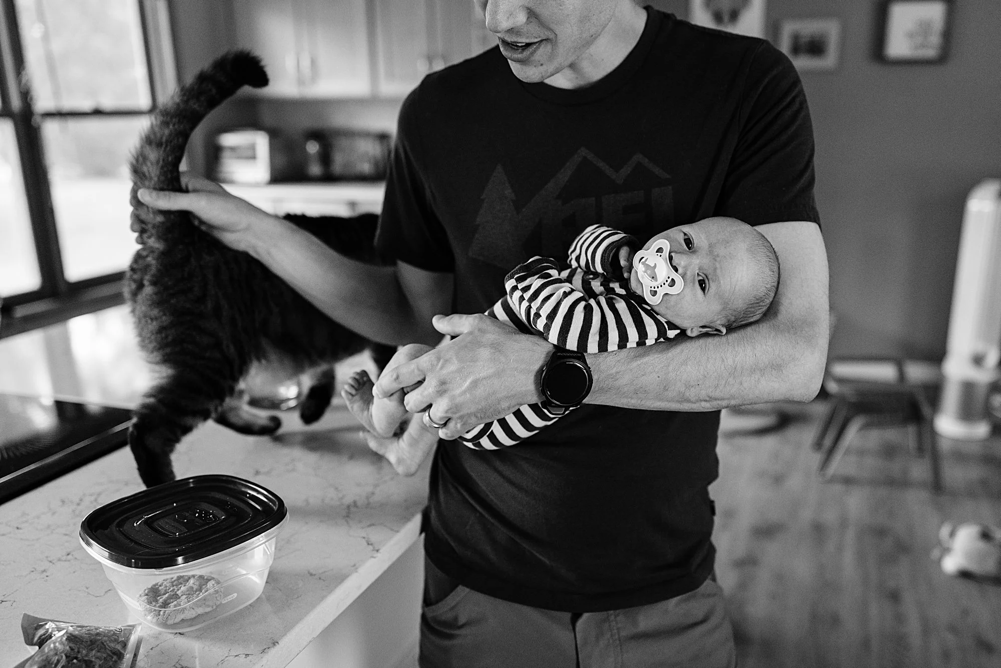 A man holding a baby with a pacifier in a striped outfit, a dark cat on the kitchen counter, and a pet food container nearby.