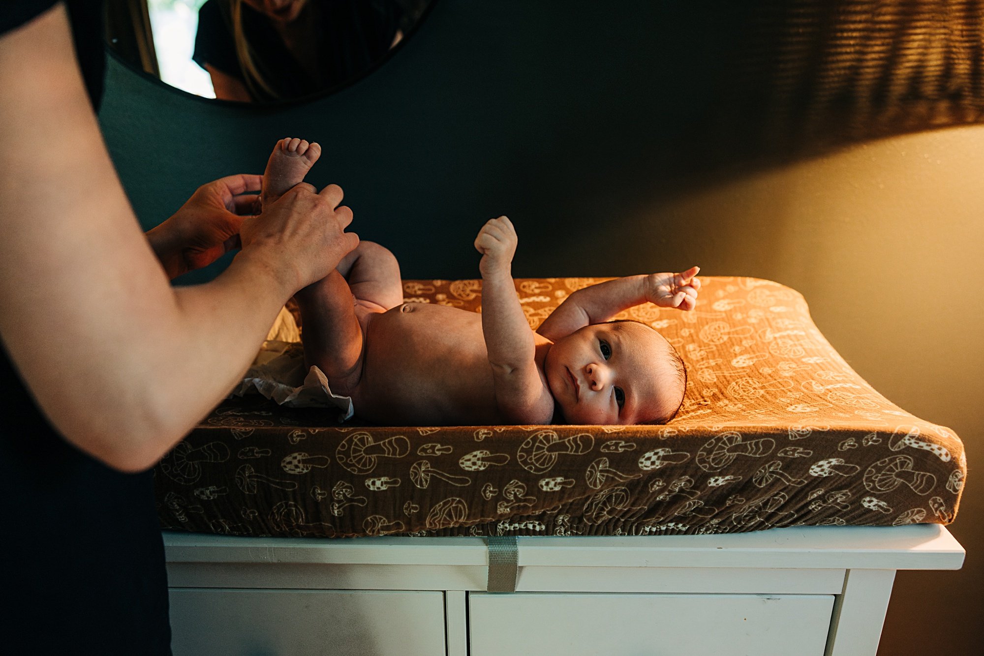 A newborn baby lying on a changing table with a patterned cover, being gently held by an adult's hands.