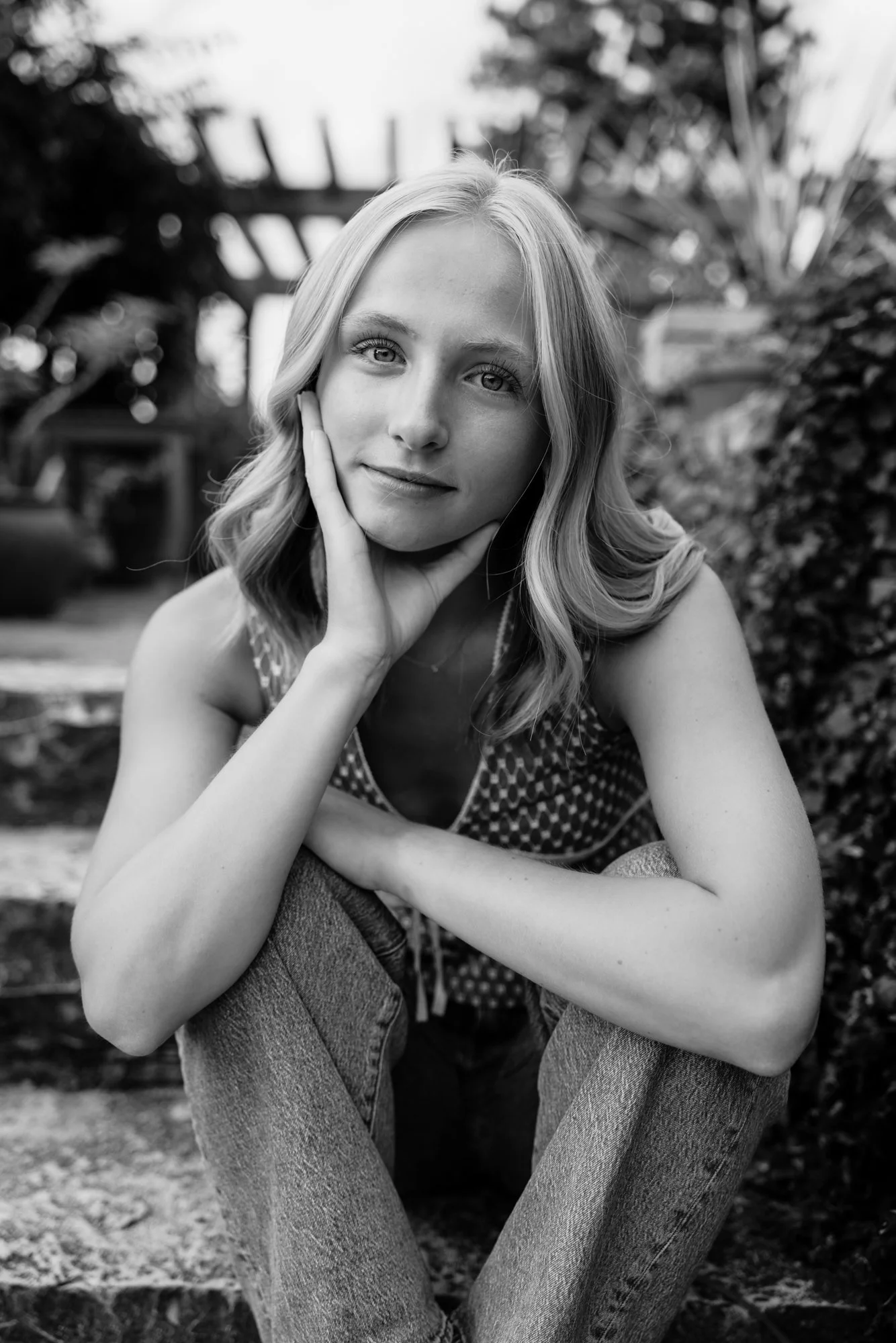 Black and white photo of a young woman with wavy hair, sitting outdoors, resting her chin on her hand, looking at the camera.