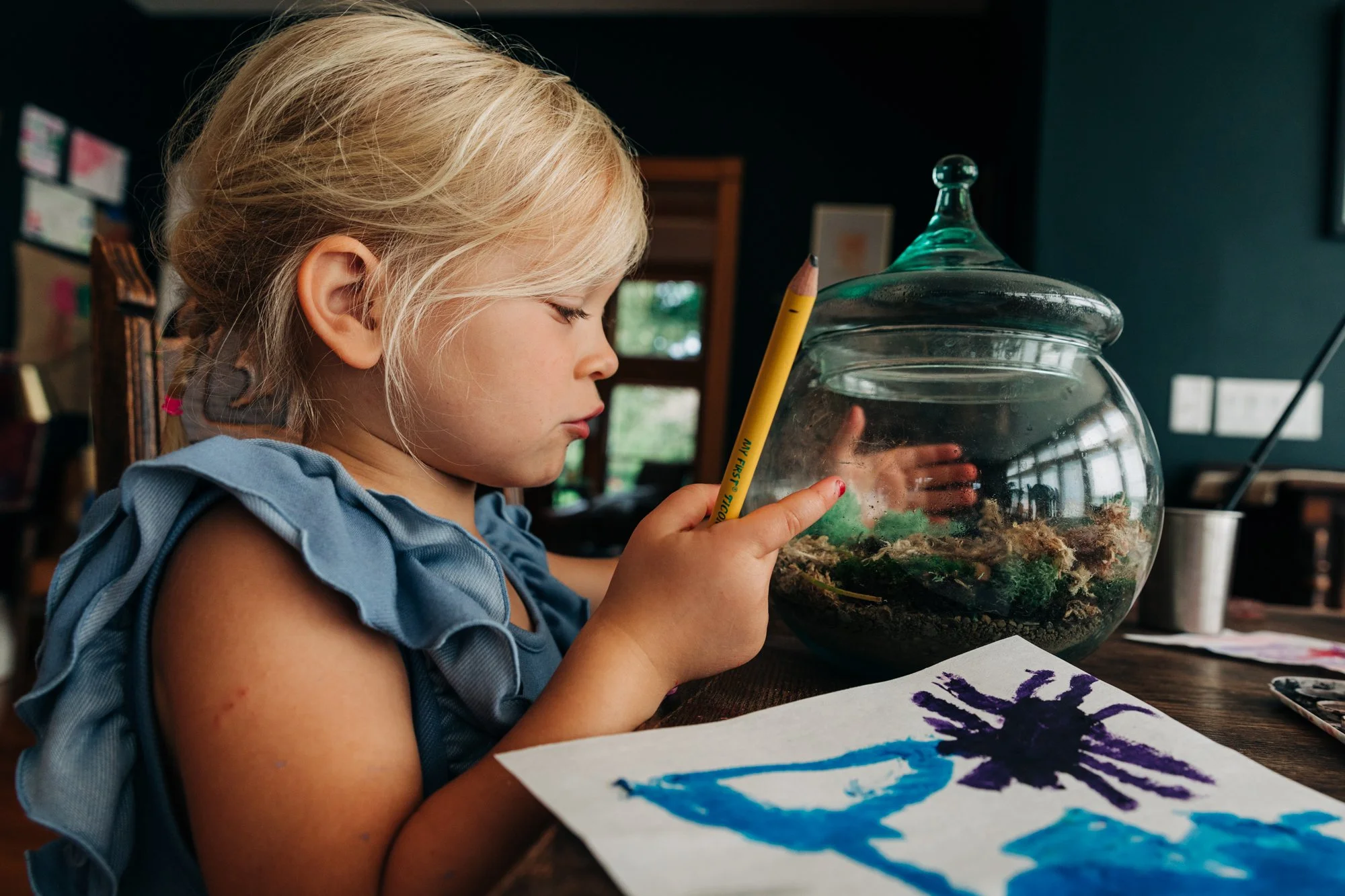 Young girl with blonde hair drawing with a sticker on her cheek, focused on her artwork at a wooden table, with a glass terrarium and painting of a spider nearby.