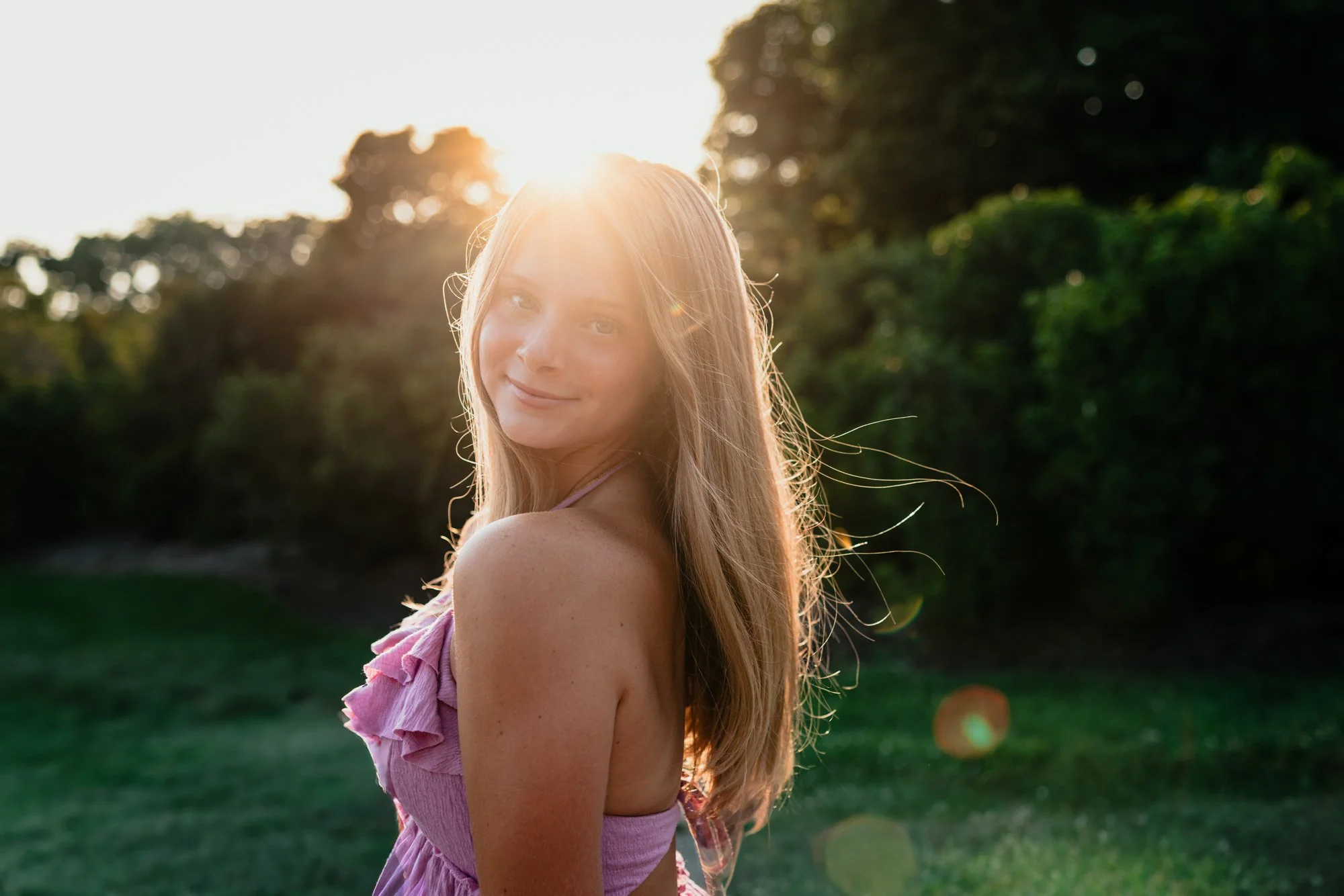 Young woman with long blonde hair wearing a pink ruffled dress outdoors during sunset, smiling at the camera. Green trees and grass are in the background.
