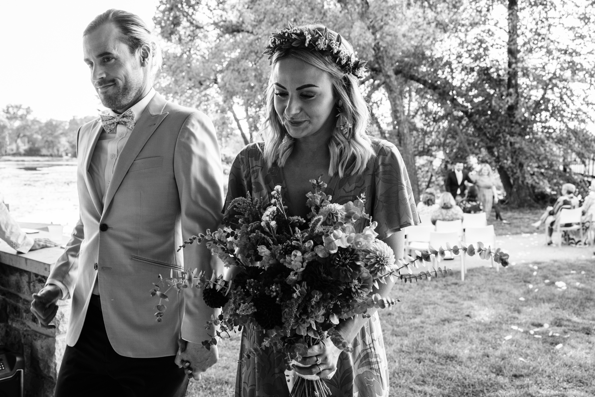 A black and white photo of a bride holding a bouquet of flowers during a wedding ceremony outdoors, with guests seated in the background.