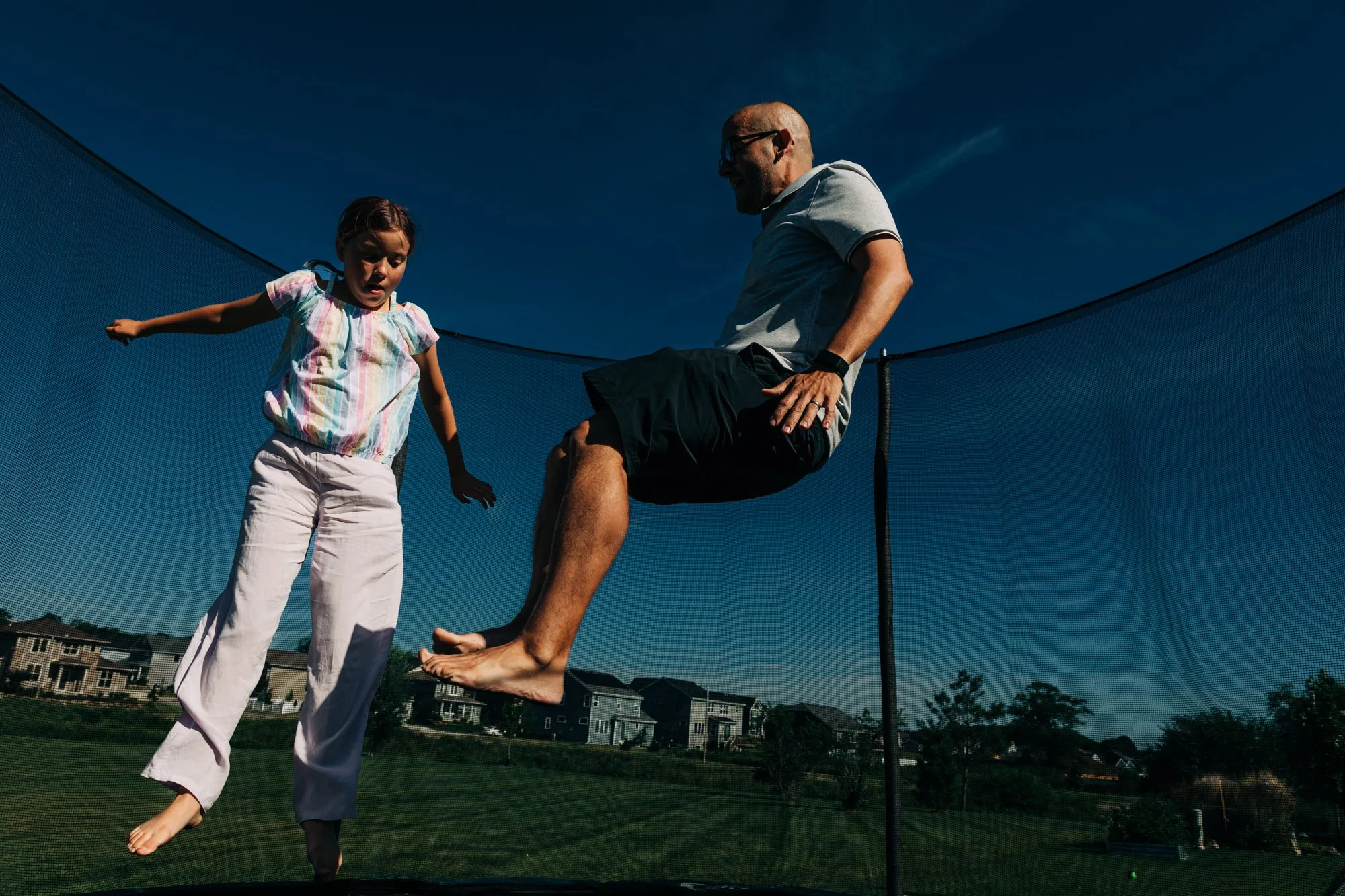 A young girl jumping on a trampoline while an older man, possibly her father, is sitting on the edge watching her.