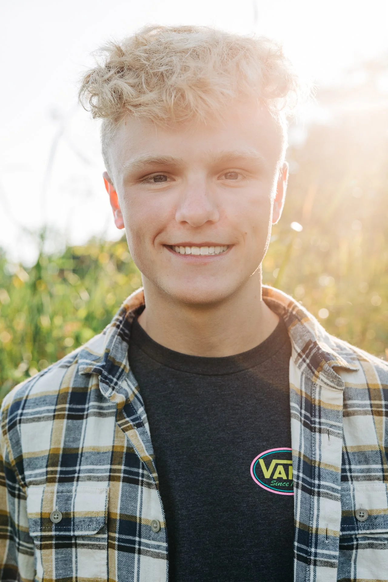 A young man with curly blonde hair and a smile, wearing a black T-shirt and a plaid shirt, standing outdoors in a field with sunlight in the background.
