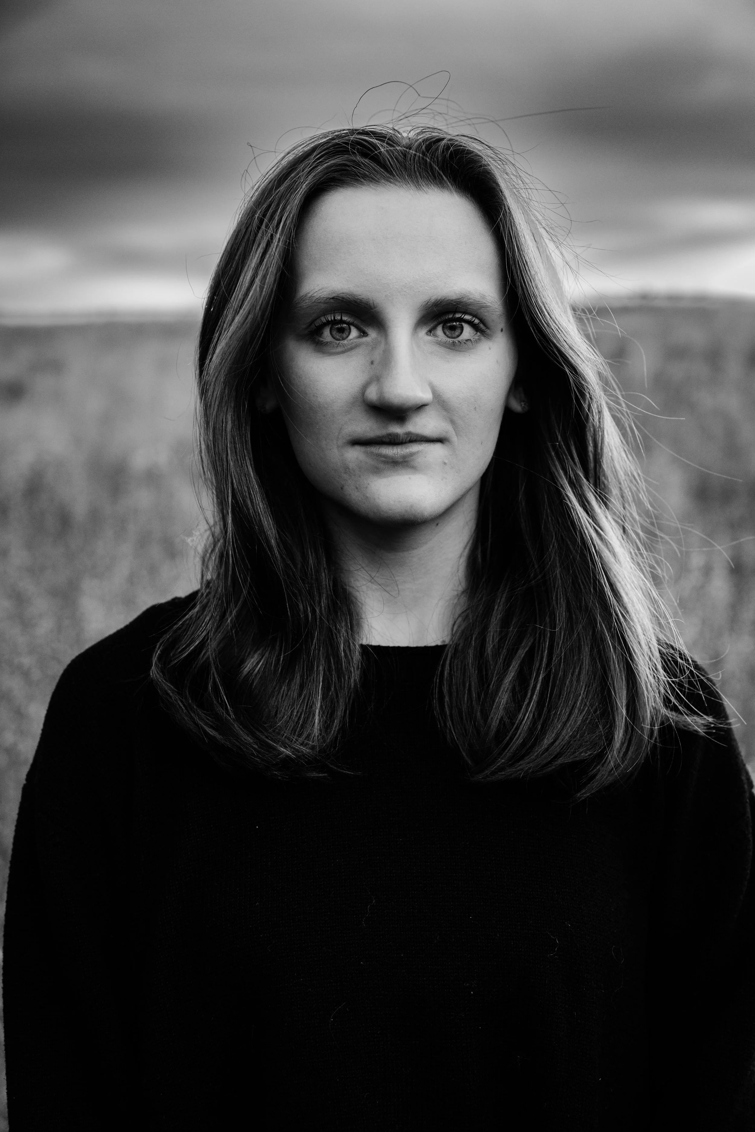 Black and white portrait of a young woman with long hair standing outdoors with a cloudy sky and field in the background.