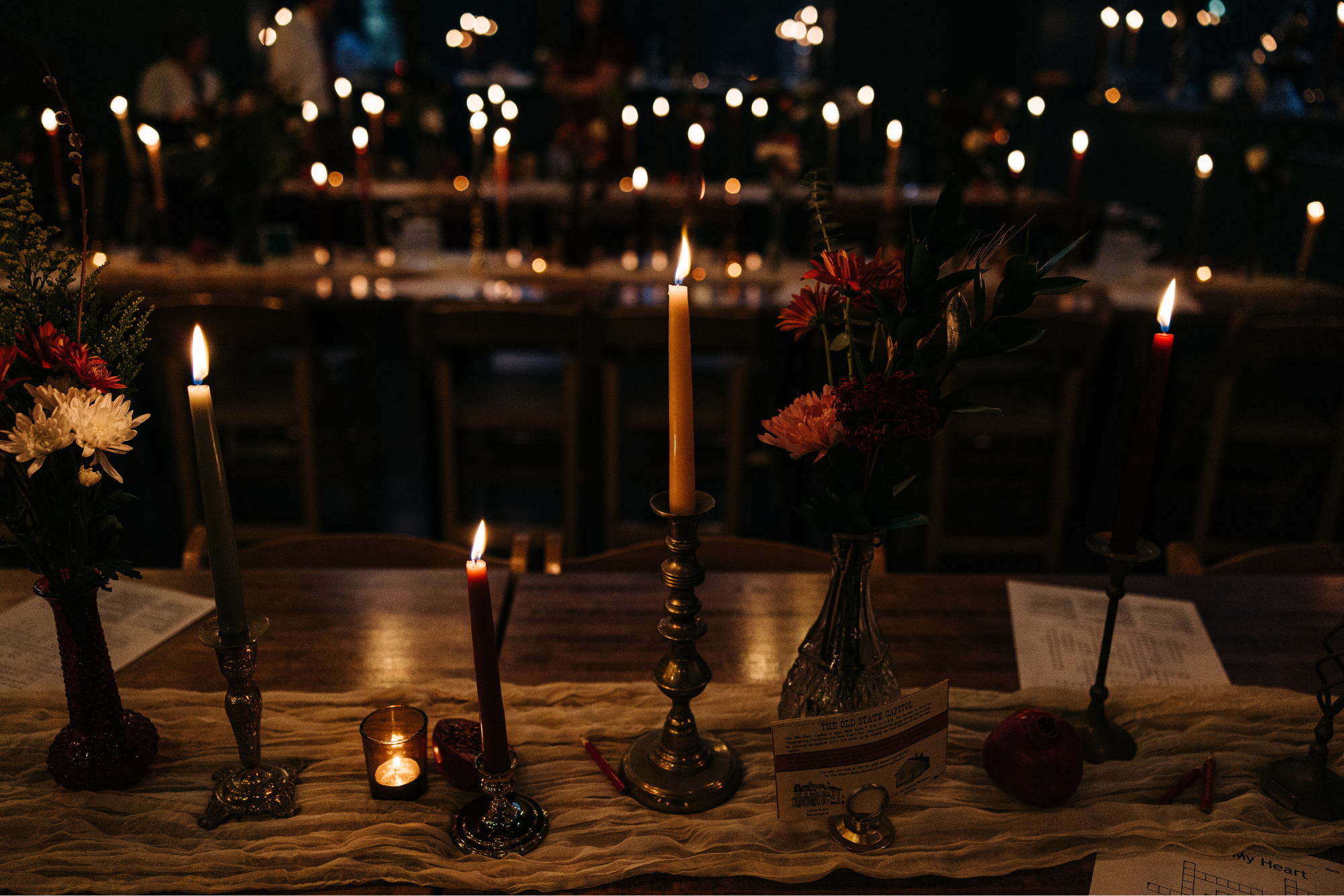 A table decorated with candles, flowers, and a small apple in a dimly lit setting, with additional candles and tables visible in the background.
