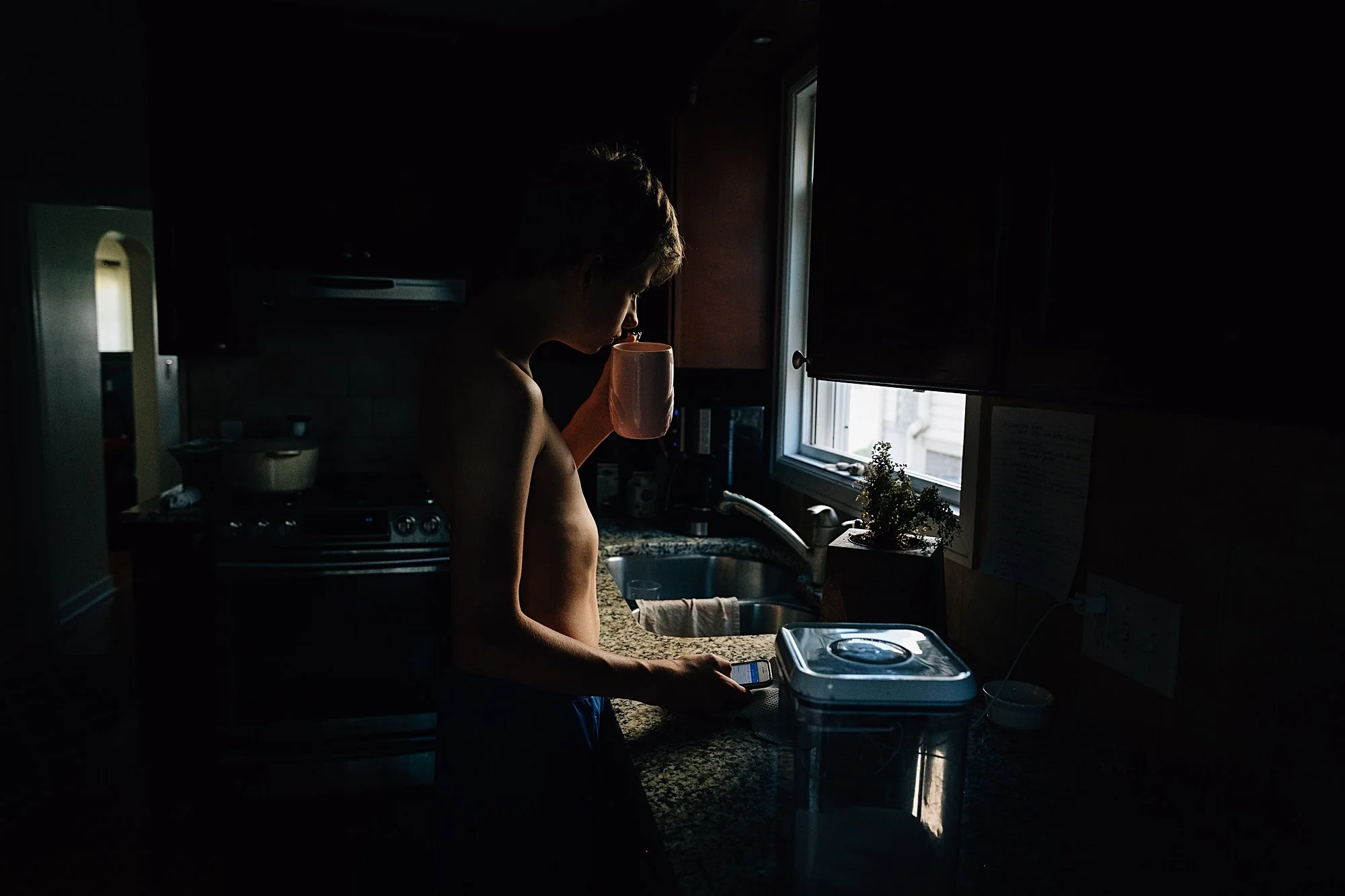 Silhouette of a young boy shirtless in a dimly lit kitchen holding a pink mug and a smartphone, with sunlight coming through a window.