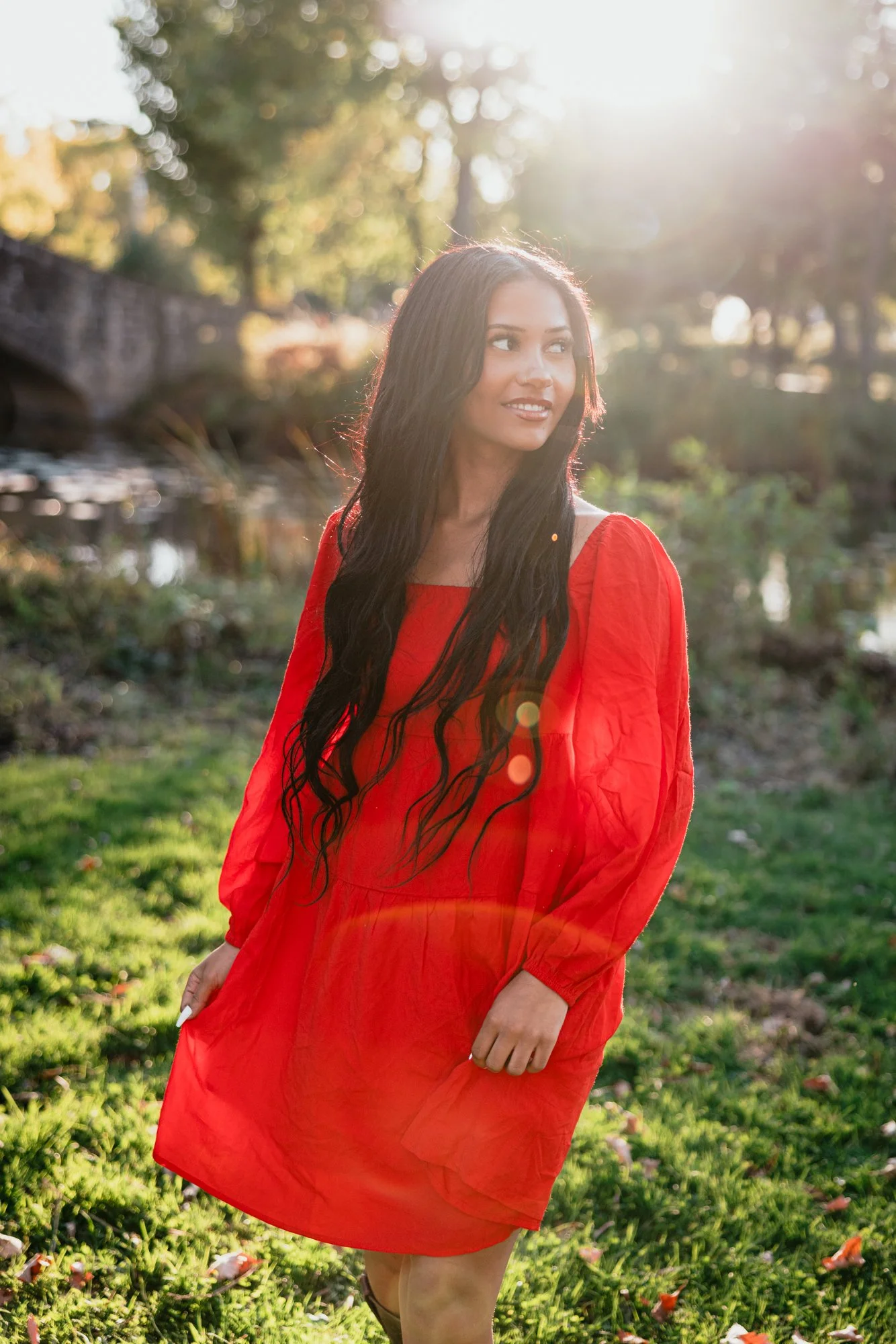 A woman with long black hair in a red dress standing outdoors in sunlight, with trees and a small bridge in the background.