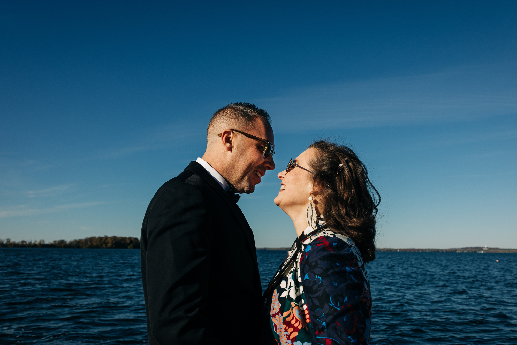 A couple, wearing glasses, is standing close by a body of water under a clear blue sky, smiling and leaning towards each other.