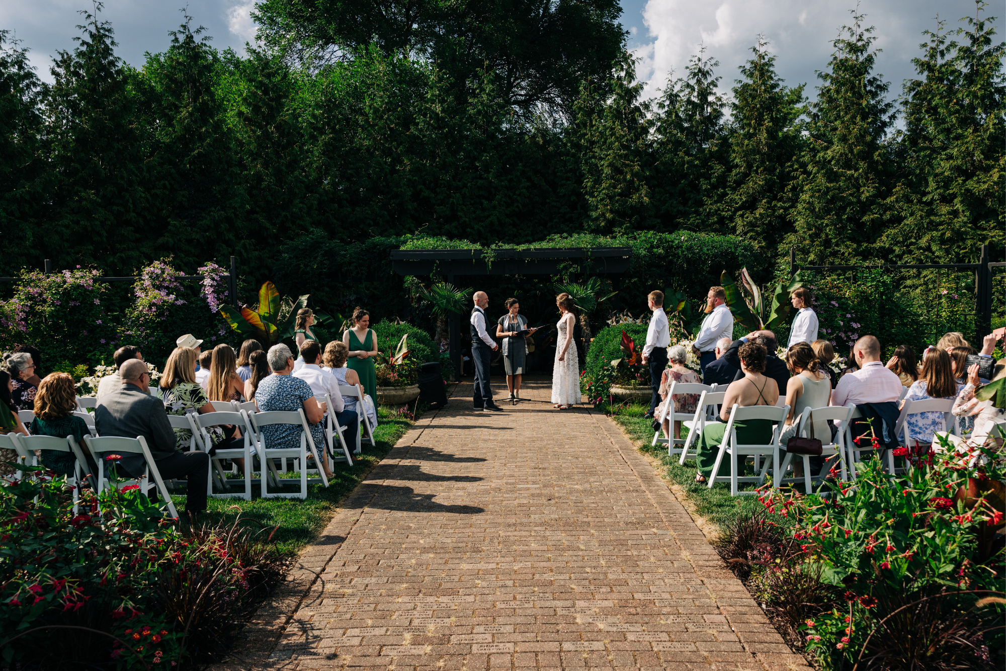 A wedding ceremony taking place outdoors in a lush green garden with trees and plants in the background. The bride and groom are standing in front of an officiant, surrounded by guests seated in white chairs on either side of a brick aisle. The atmos