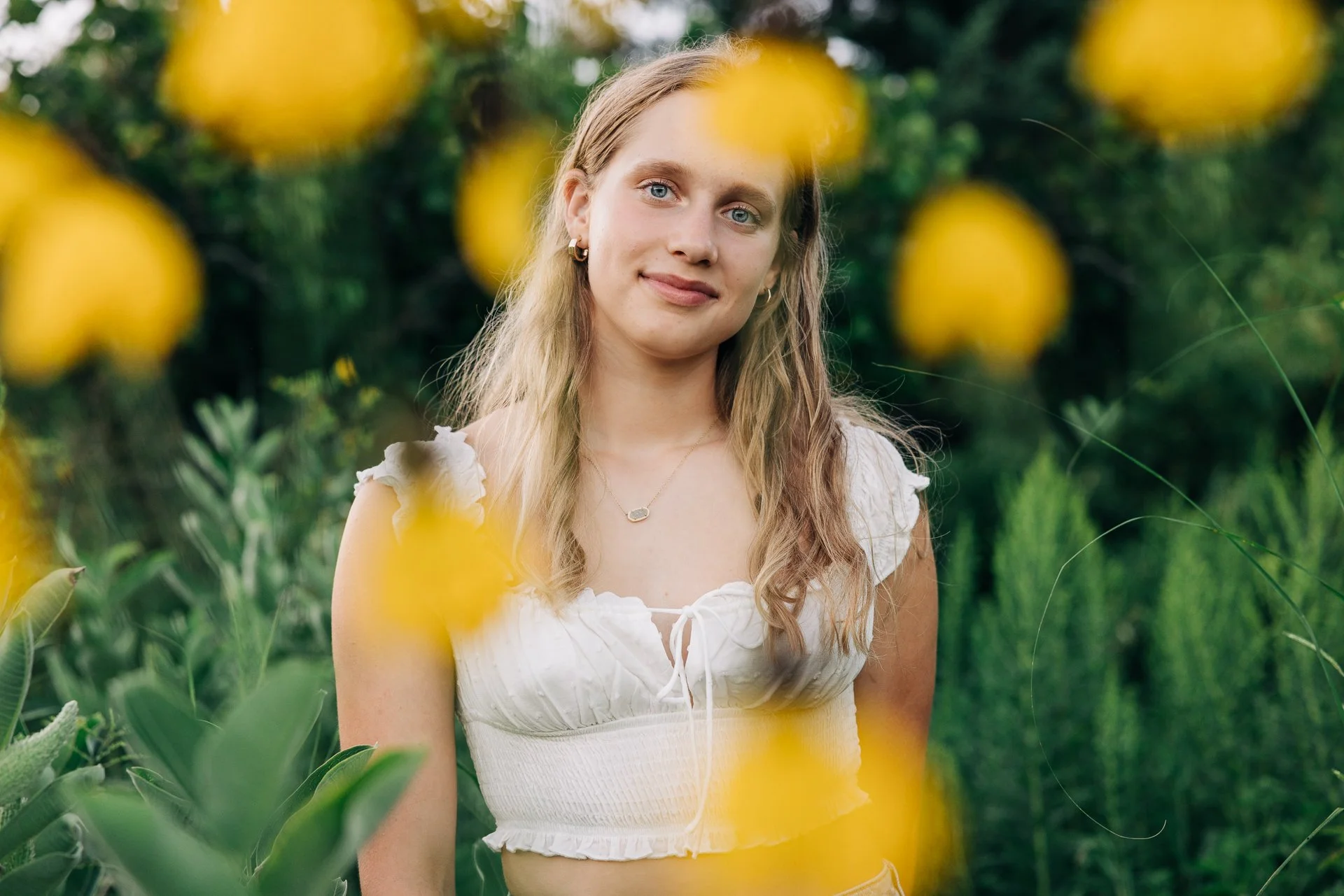 A young woman with long blonde hair, wearing a white ruffled crop top, standing outdoors among greenery and yellow flowers.
