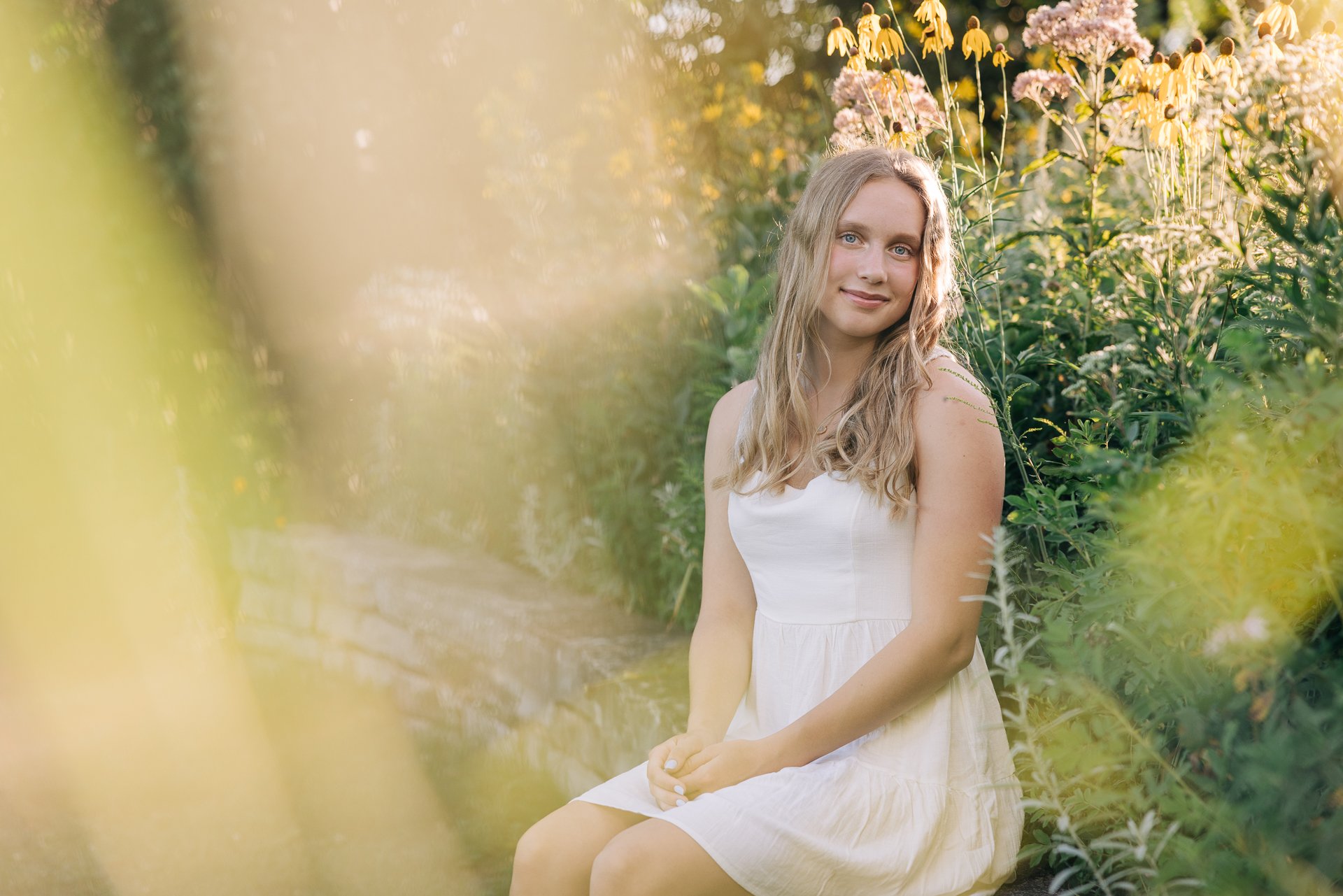 A young woman with long wavy hair sitting outdoors on a stone ledge, wearing a white dress, surrounded by plants and flowers with sunlight in the background.