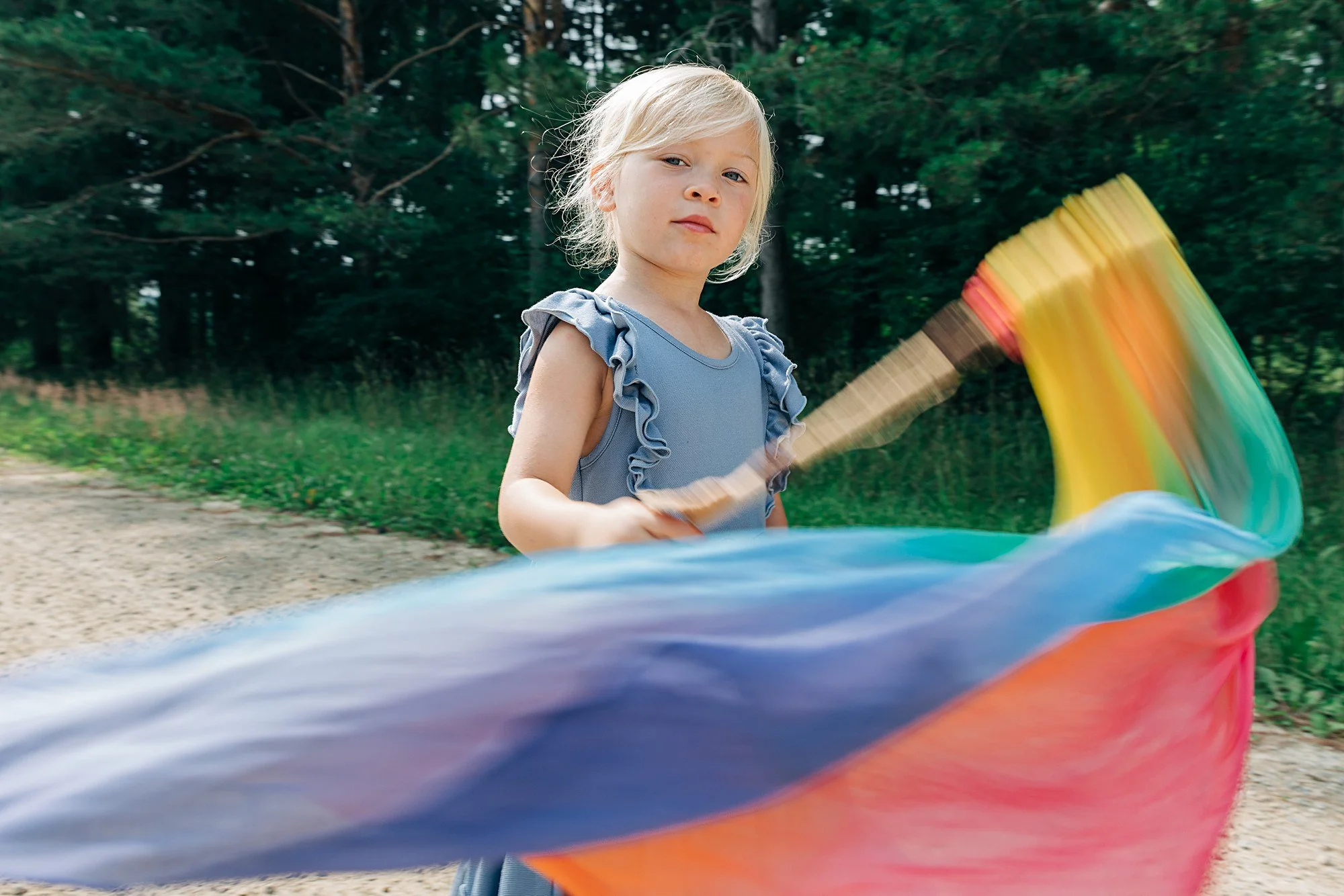 A young girl with blonde hair wearing a blue ruffled sleeveless top spinning a colorful rainbow flag outdoors near a wooded area.