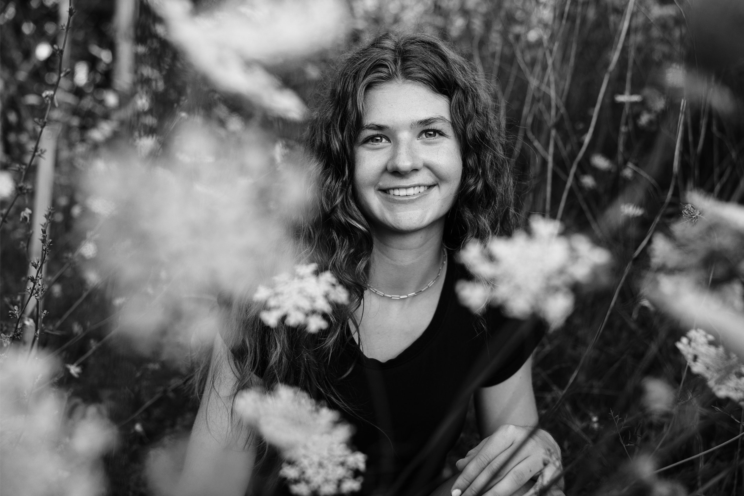 Black and white portrait of a young woman with curly hair, smiling, surrounded by blurred flowers and plants in outdoor setting.