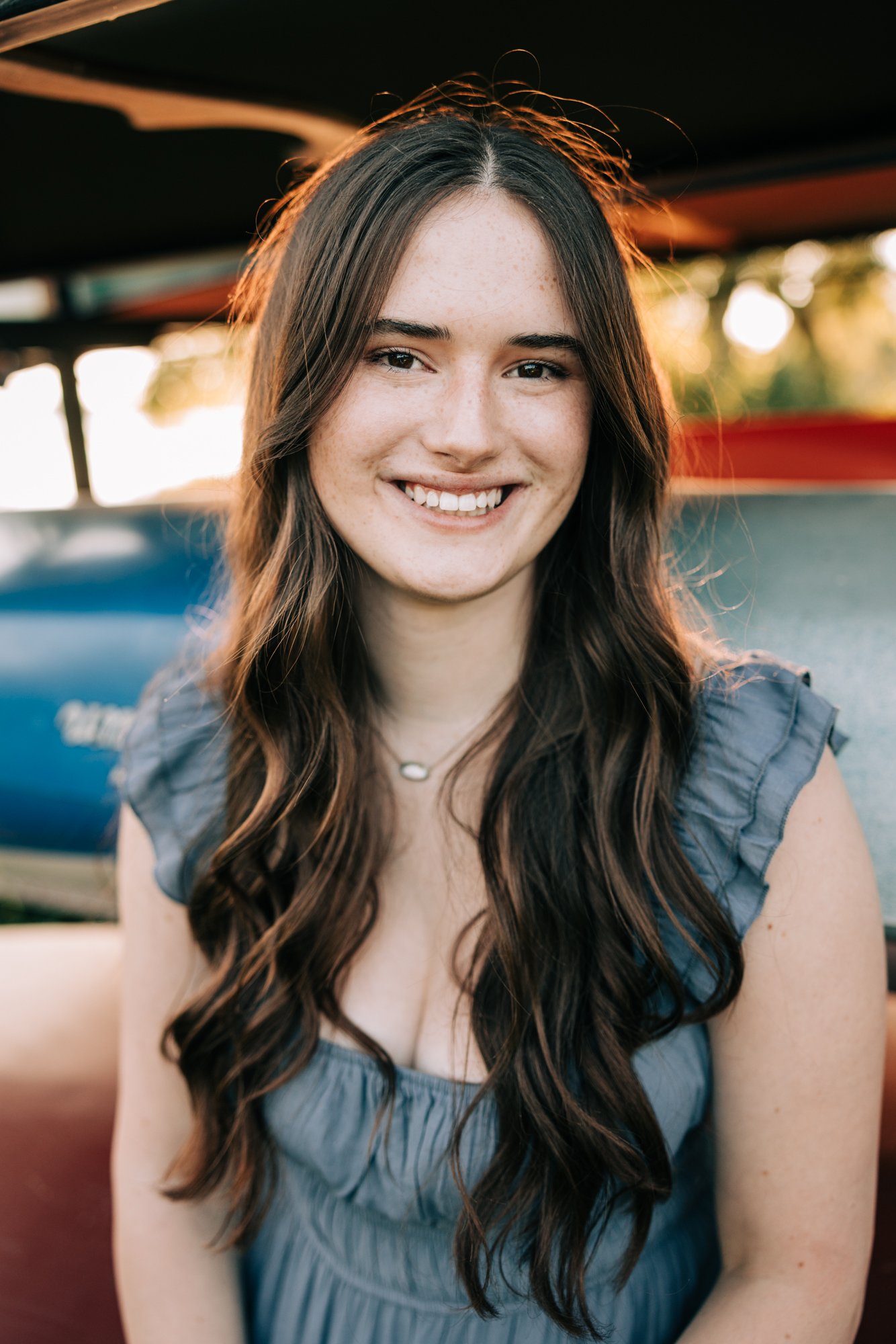 A smiling young woman with long, wavy brown hair outdoors during sunset. She is wearing a ruffled blue dress and a necklace. The background is blurred with some boats and greenery.