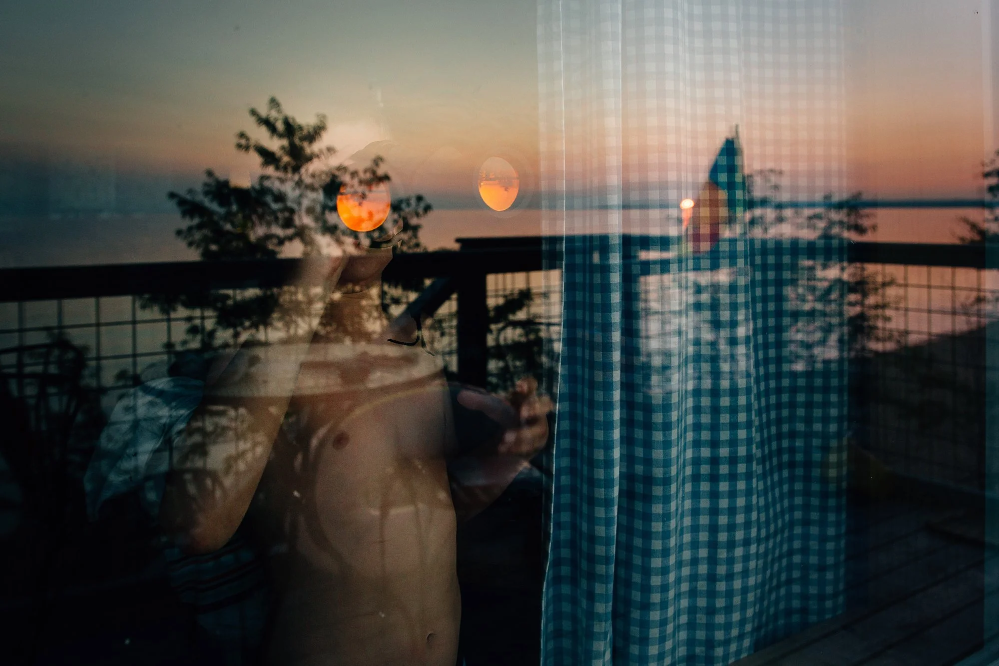 Reflection of a child in a window at sunset, with trees and a blue checkered curtain visible.