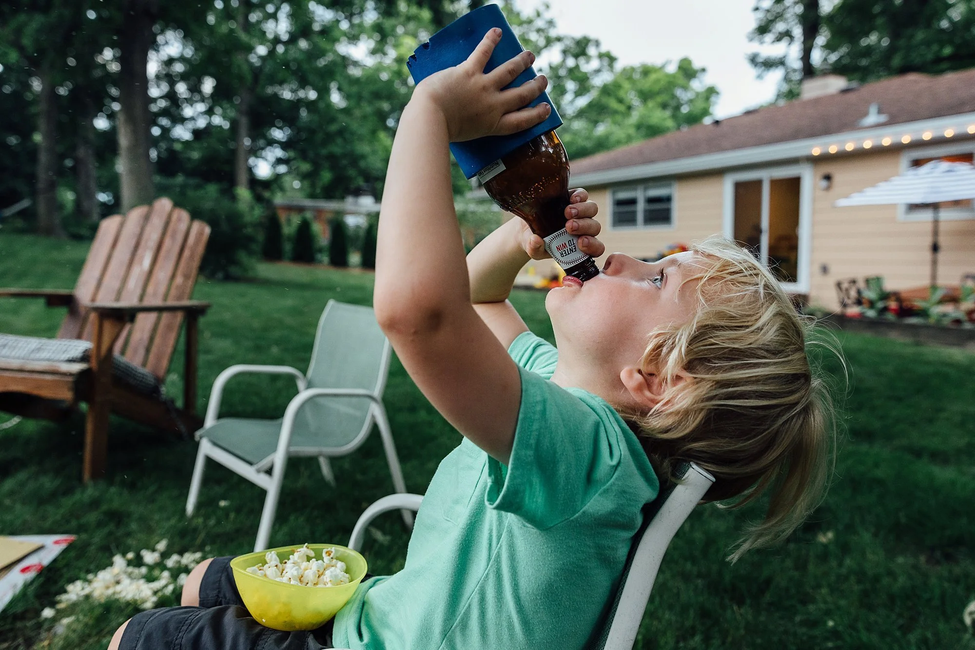 A young boy with blonde hair sitting outdoors in a backyard, drinking from a brown glass bottle held upside down, with a yellow bowl of popcorn on his lap.