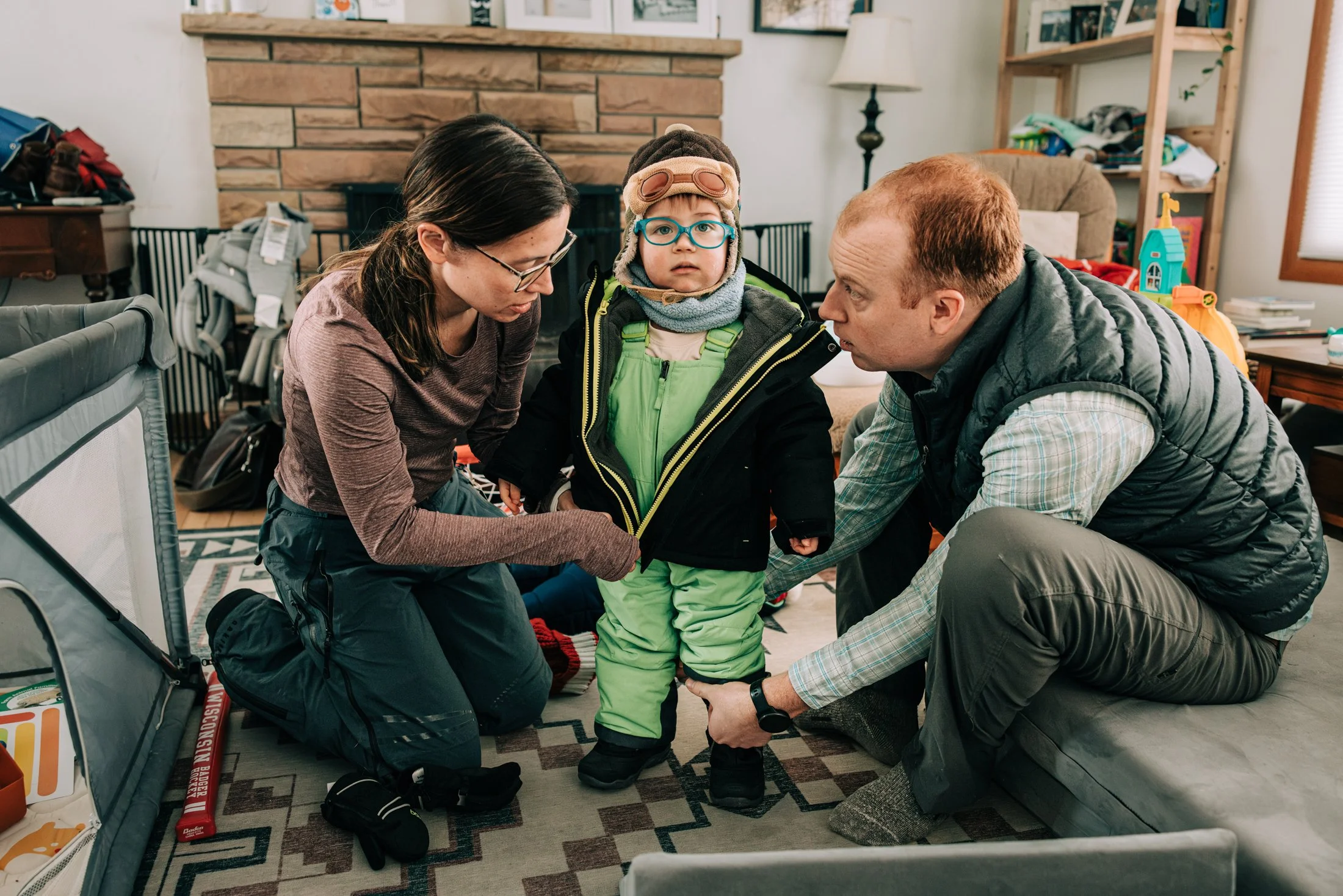 A young child dressed in a winter jacket and snow gear stands between two adults in a living room, with one adult measuring the child's leg.