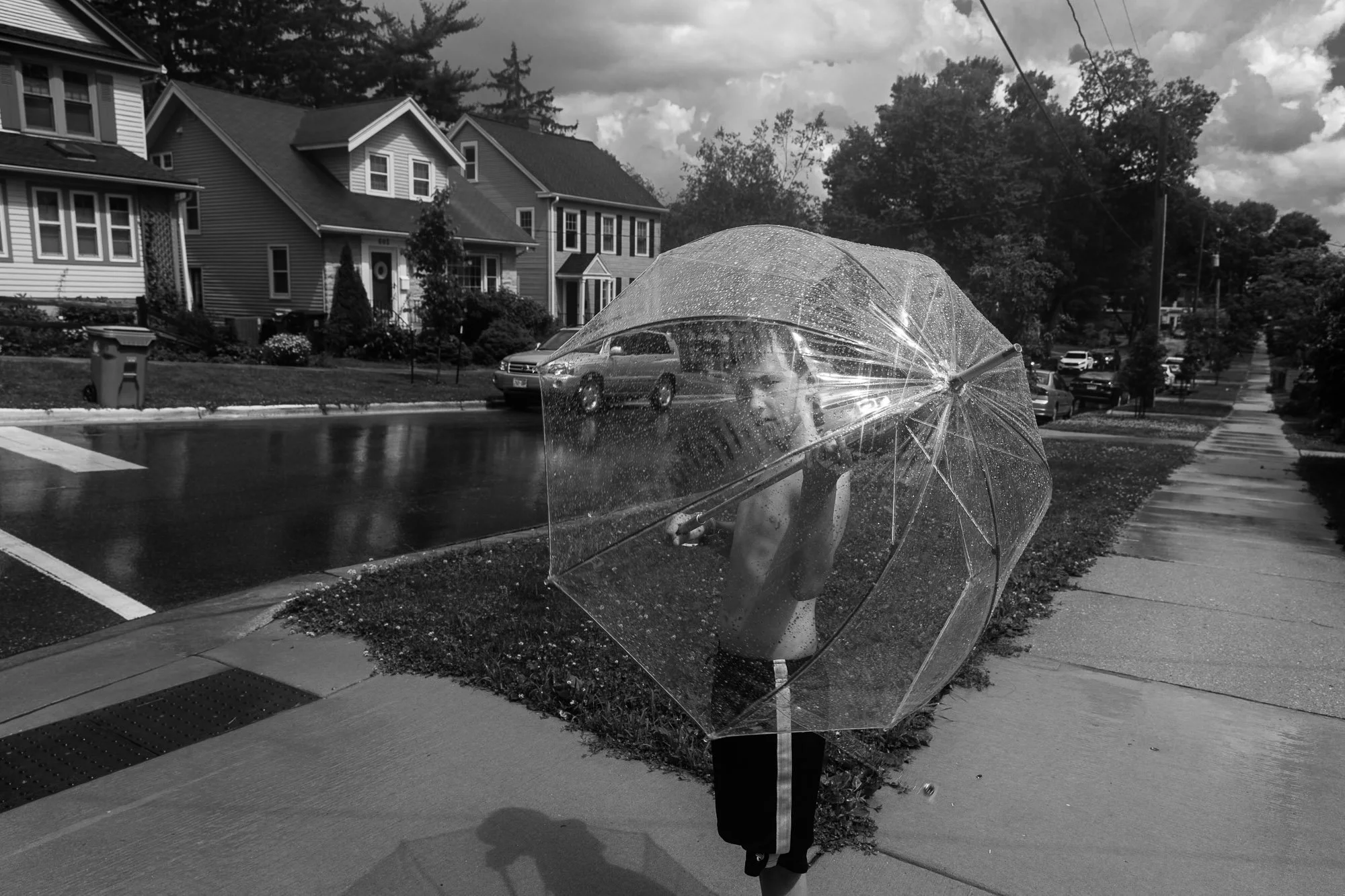 A young boy holding a transparent umbrella on a rainy day in an urban neighborhood, with wet pavement and parked cars visible.